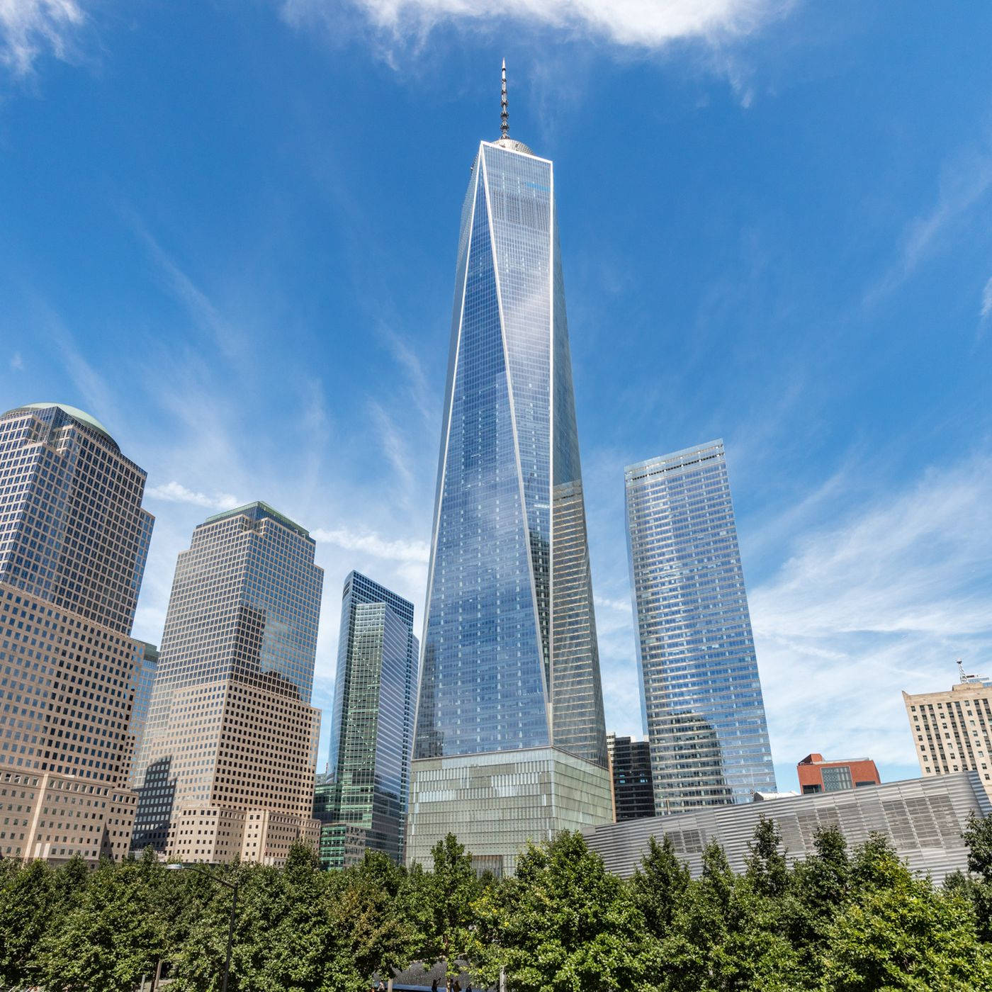 Building 桌布 壁紙 — 一座高聳的玻璃摩天大樓刺穿明亮的藍天，帶有縷縷白雲，周圍環繞著其他現代建築和其底部的綠色樹木。
