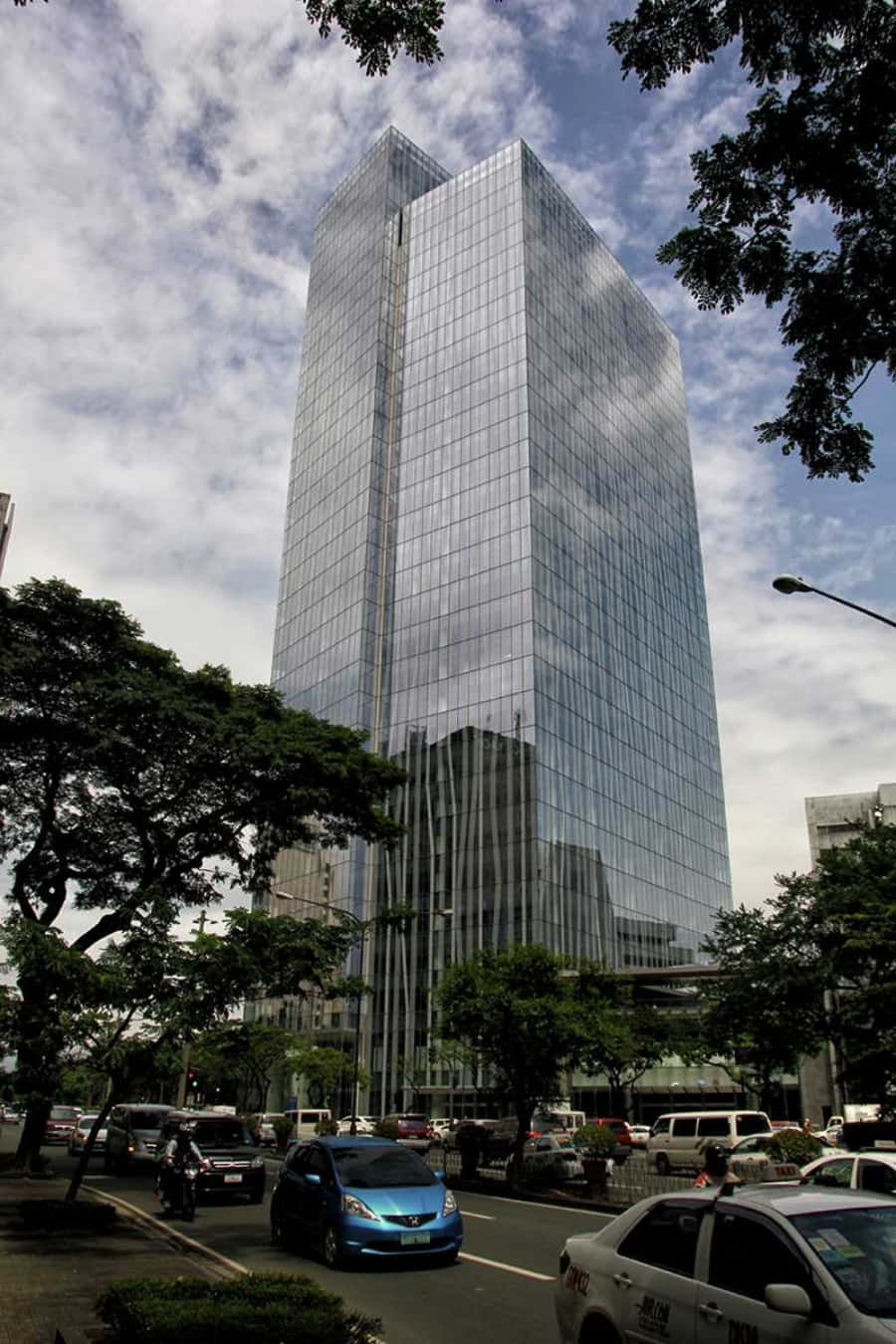 Building 桌布 壁紙 — 一座高聳的玻璃摩天大樓反射著多雲的天空，汽車和樹木環繞其現代都市的底座。