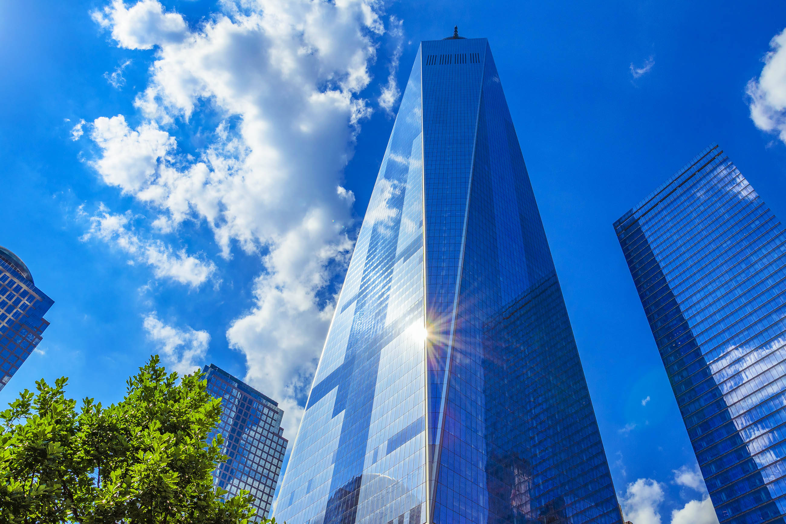 Building 桌布 壁紙 — 一座高聳的玻璃摩天大樓在湛藍的天空、蓬鬆的白雲和其底部的綠樹映襯下反射著陽光。