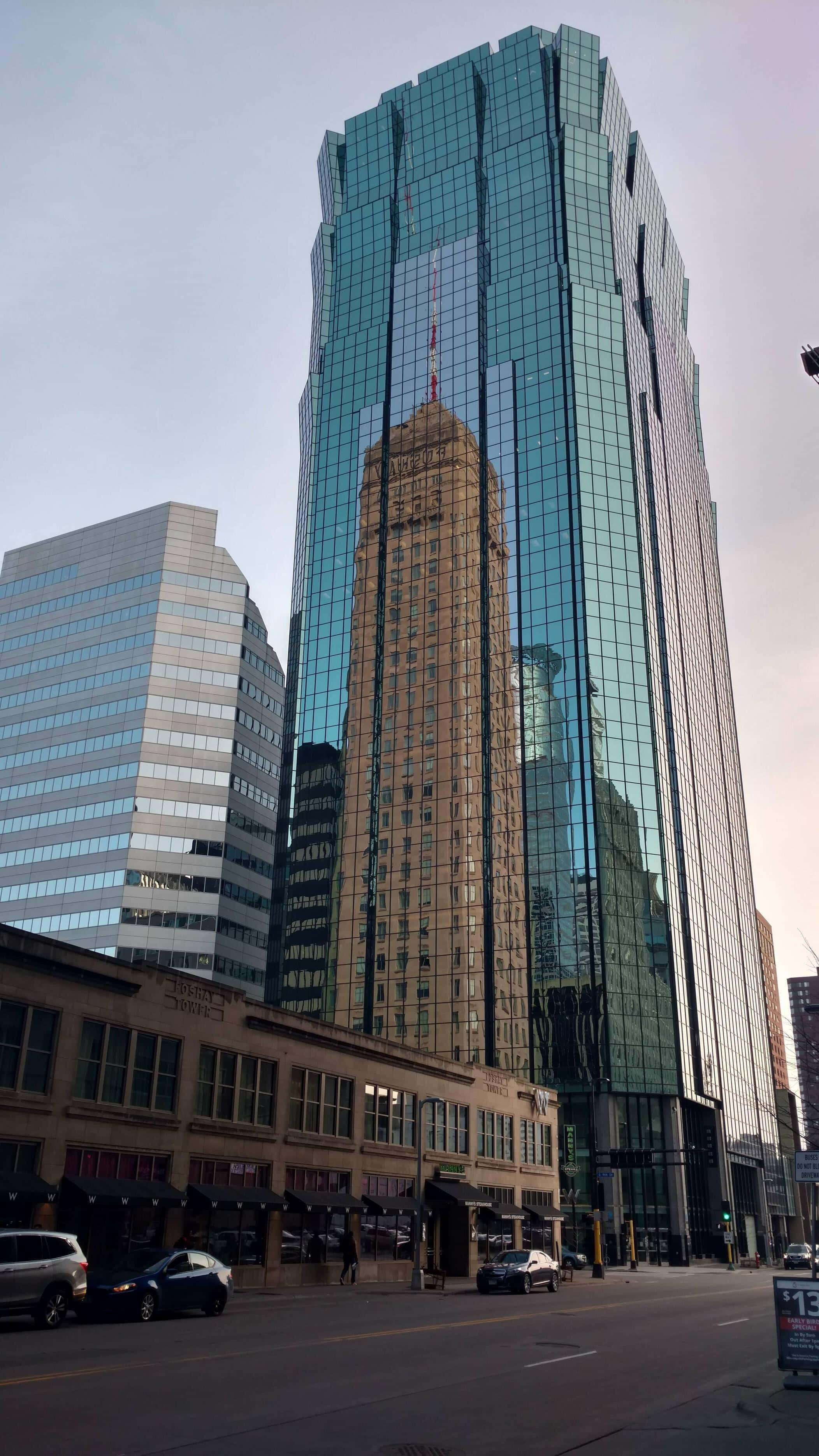 Building 桌布 壁紙 — 一座高聳的玻璃摩天大樓在繁忙的城市街道場景中，映照著一棟歷史建築，背景是蒼白的天空。