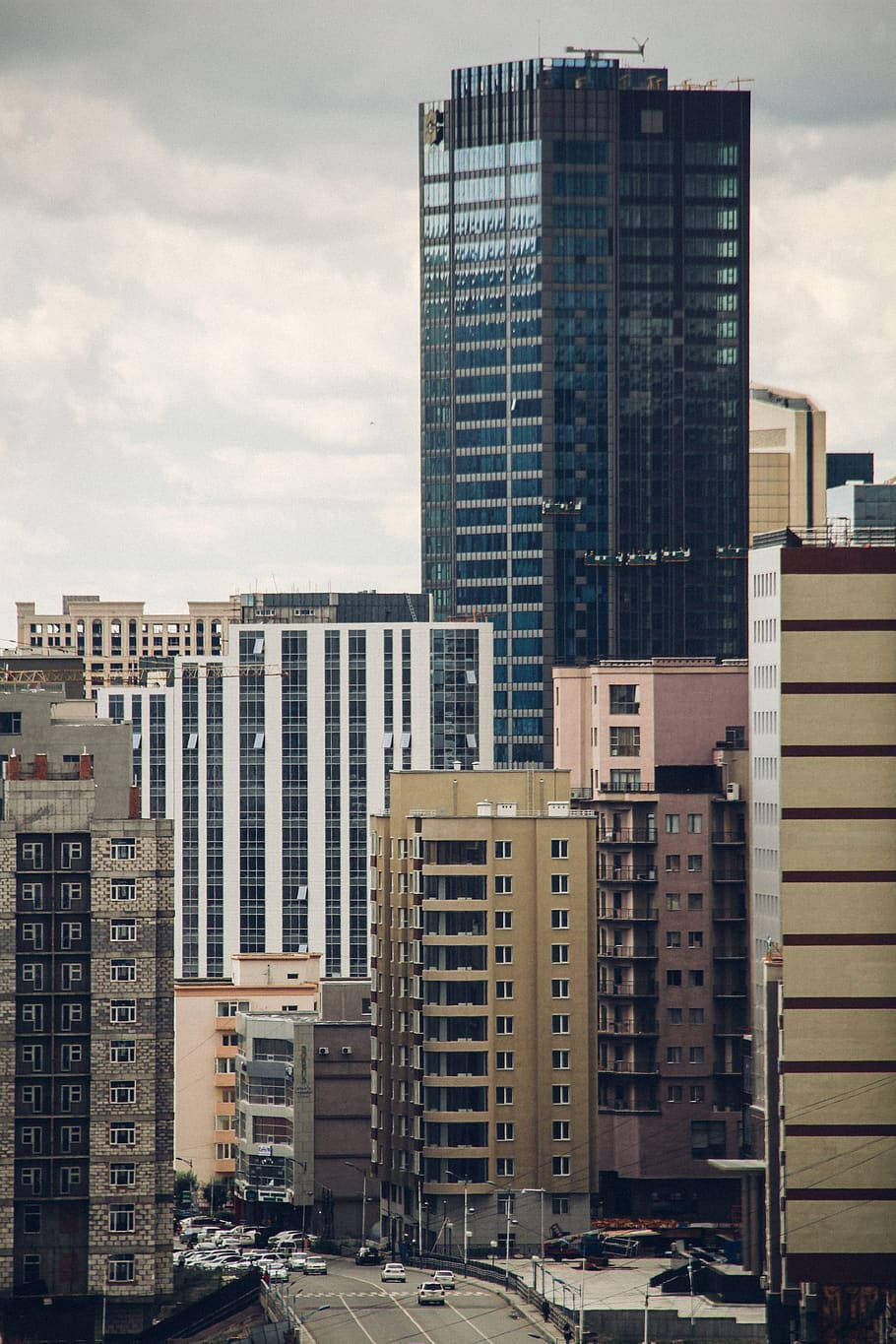Buildings 桌布 壁紙 — 一座高聳的玻璃摩天大樓在陰天之下主導著密集的城市天際線，周圍有各種建築，下方是街道。