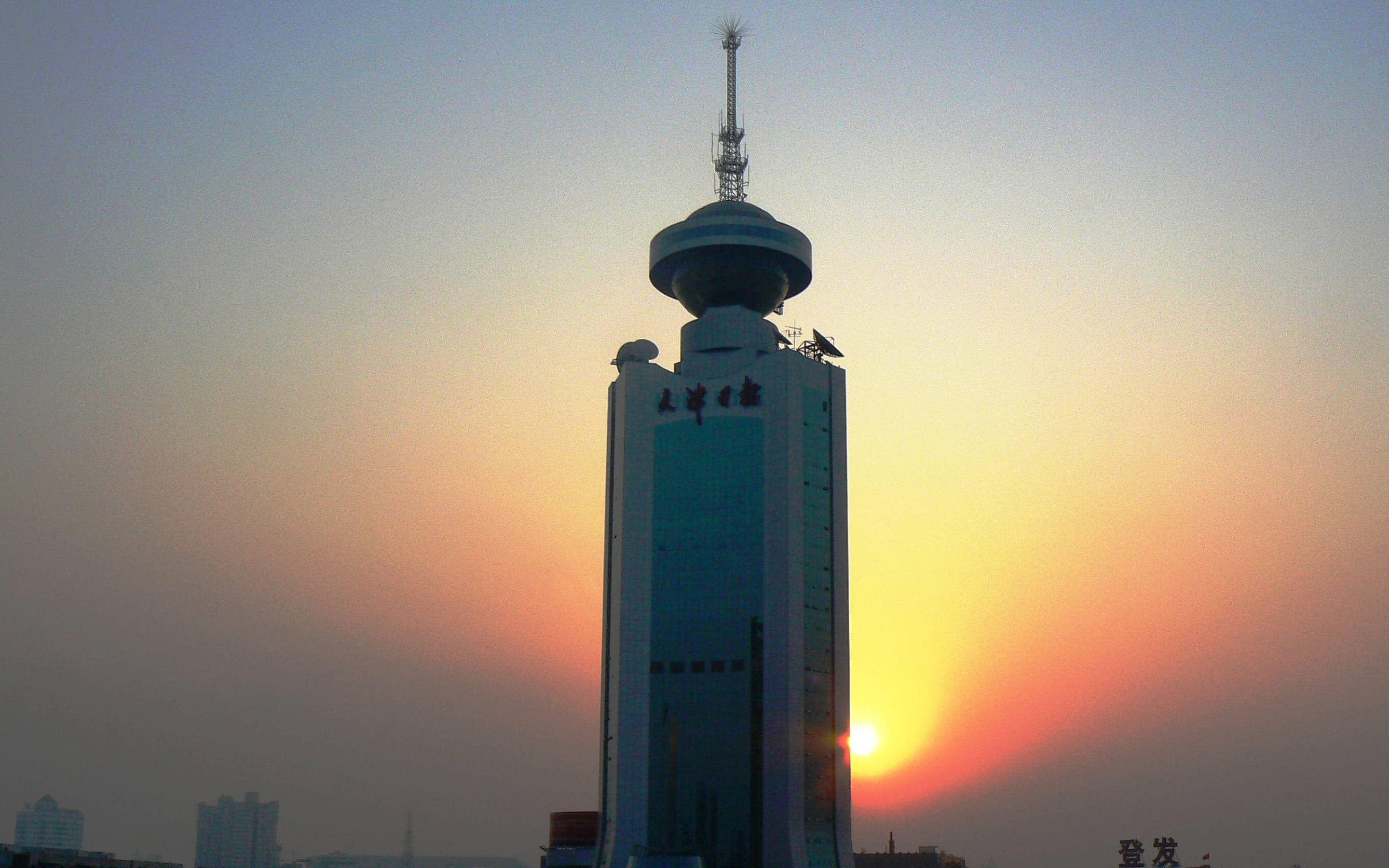 Tianjin 桌布 壁紙 — 一座高聳的現代塔樓在溫暖的橙色和粉紅色調的鮮豔日落天空下形成剪影。