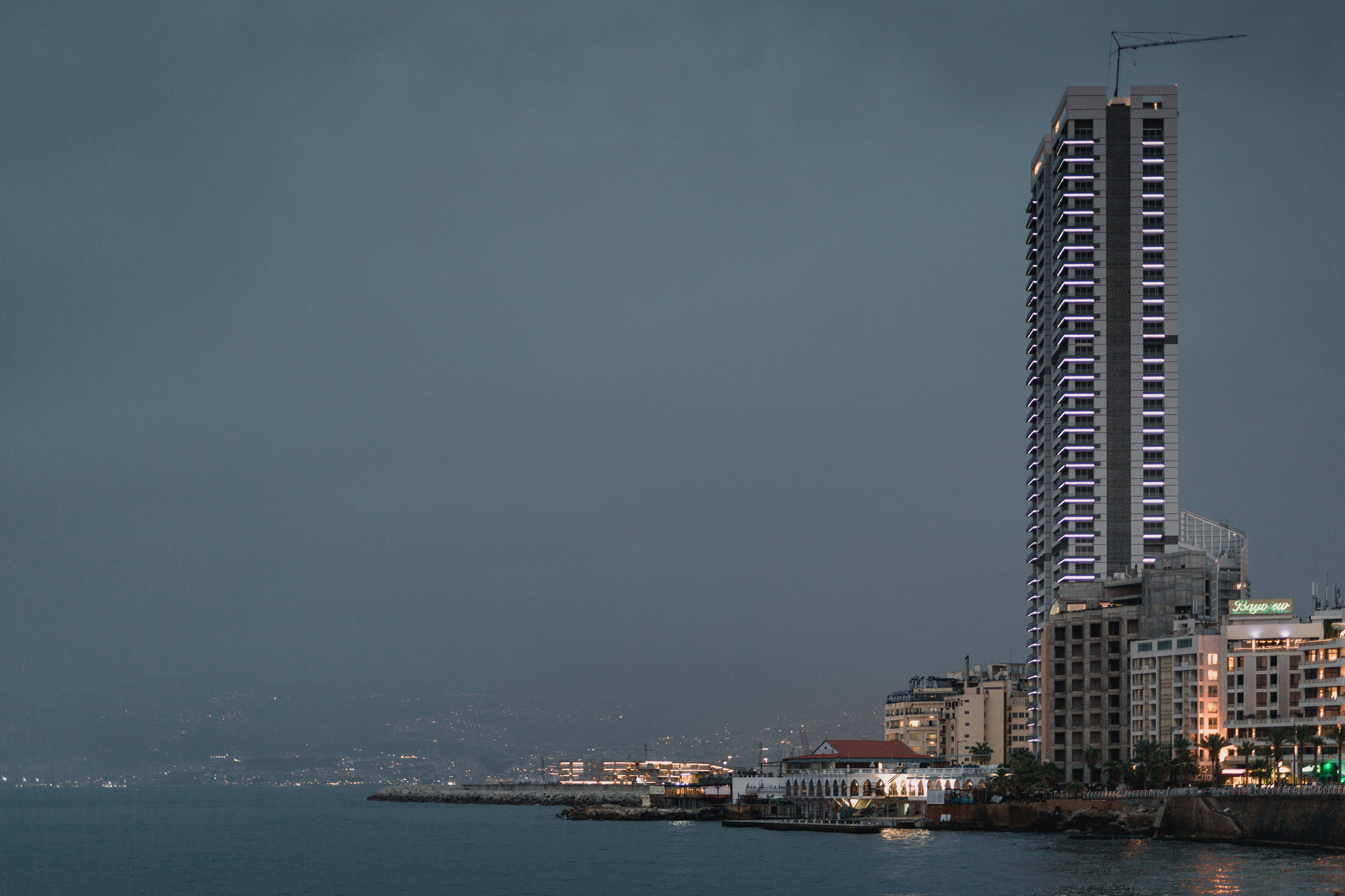 Lebanon 桌布 壁紙 — 一座高聳的現代摩天大樓，帶有紫色燈光，在黃昏時分矗立在陰暗多雲的天空下，旁邊是平靜的海濱。