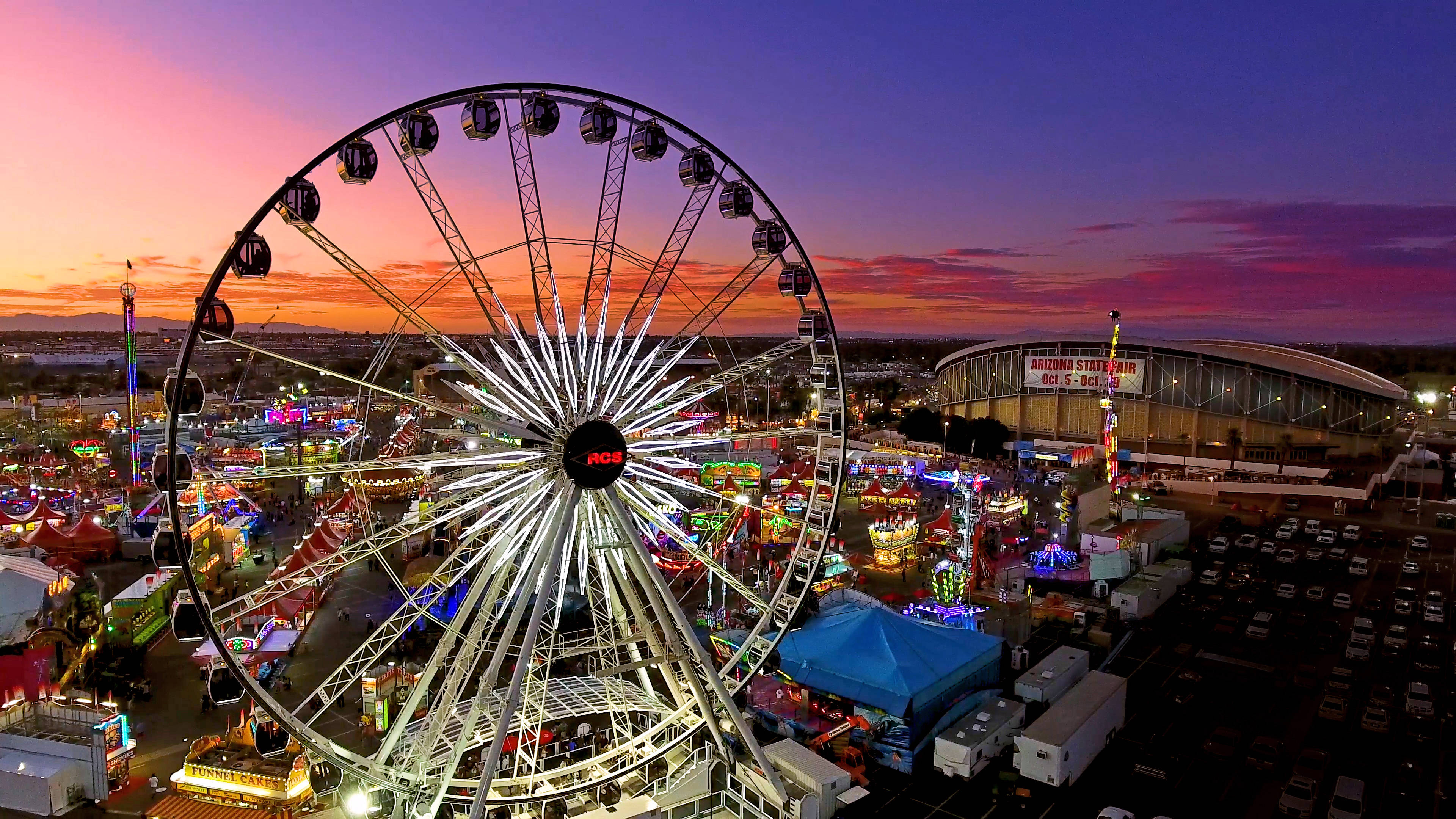 日落 桌布 壁紙 — 一座高聳的發光摩天輪在暮色中主宰著熱鬧的嘉年華，色彩繽紛的遊樂設施和閃爍的燈光映襯著紫橙色的天空。