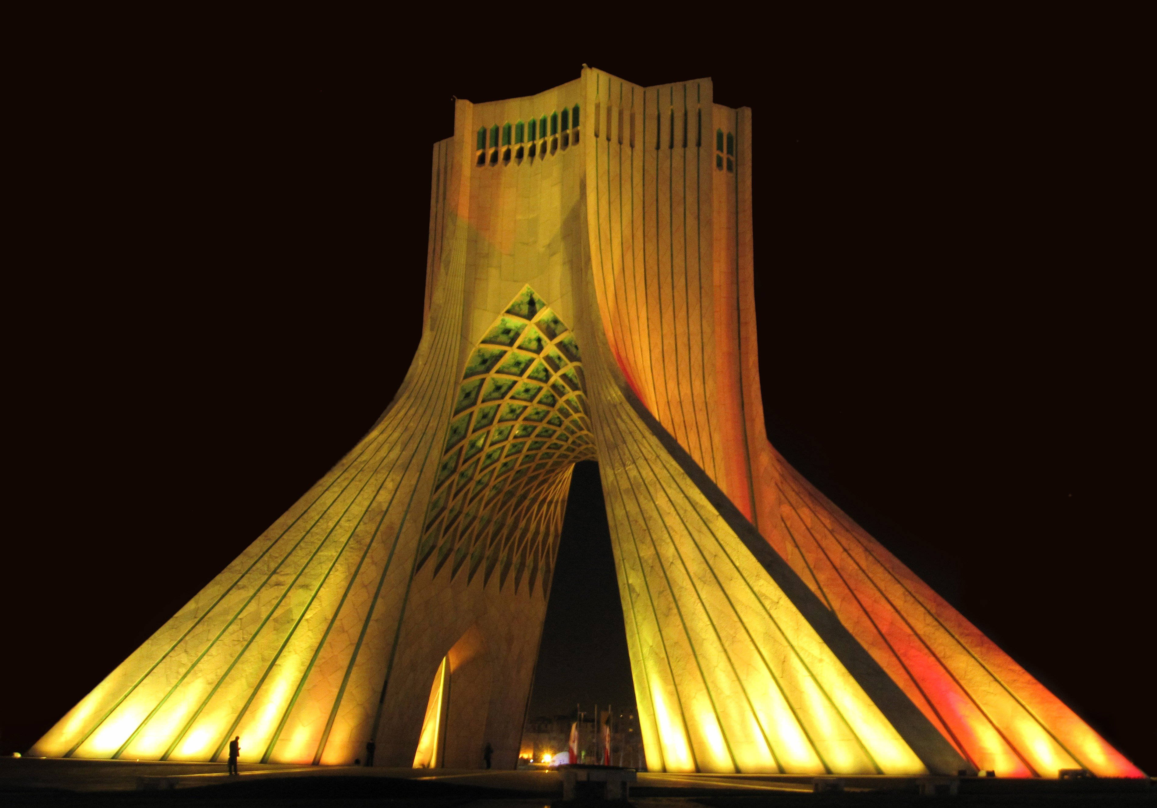 Tehran 桌布 壁紙 — 一座高聳的發光紀念碑，帶有幾何圖案，在黑暗的夜空中溫暖地發光。