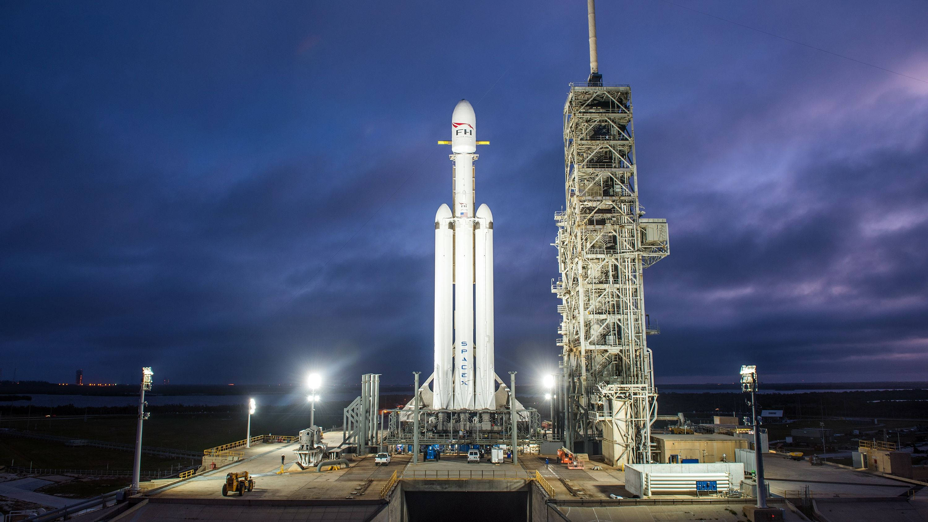 SpaceX 桌布 壁紙 — 一座高聳的白色SpaceX火箭在黃昏時分被照亮，矗立在發射塔旁，背景是戲劇性的紫色天空。
