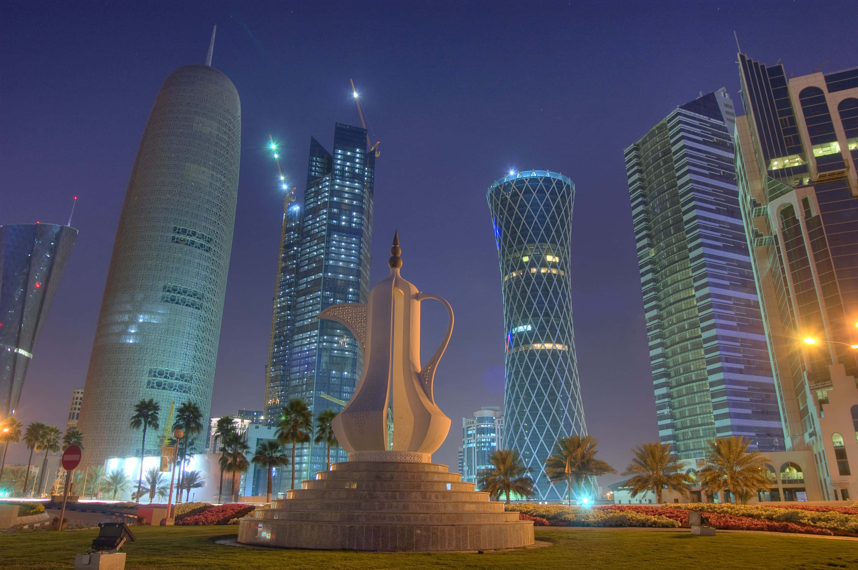 Qatar 桌布 壁紙 — 一座高聳的白色咖啡壺雕塑矗立在夜間整潔的公園中，被發光的未來主義摩天大樓和棕櫚樹環繞，背景是深藍色的天空。