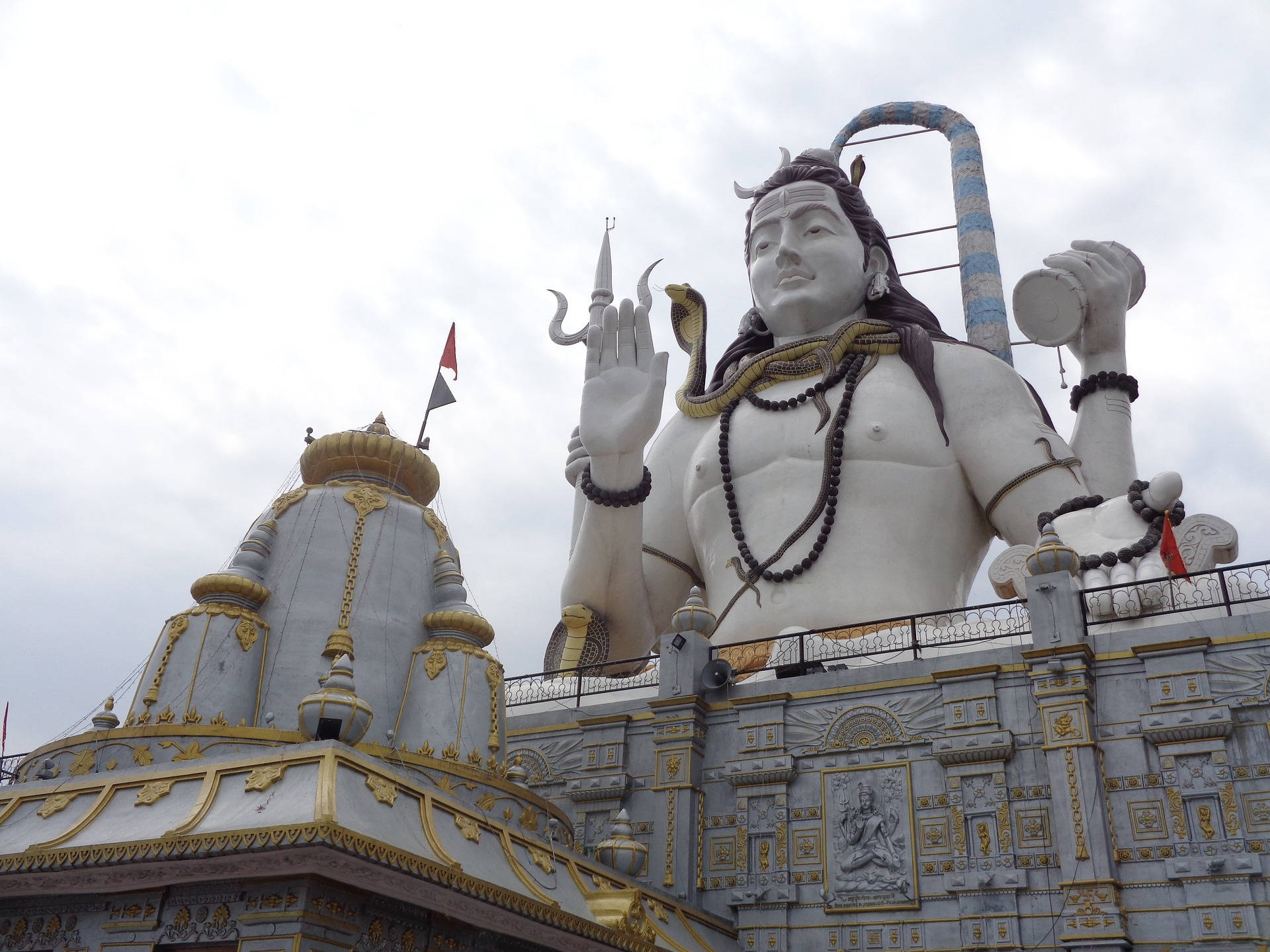 shiv shankar 桌布 壁紙 — 一座高聳的白色濕婆神像，戴著眼鏡蛇項鍊，手持三叉戟，矗立於一座飾有金色點綴的華麗寺廟頂端，天空多雲。