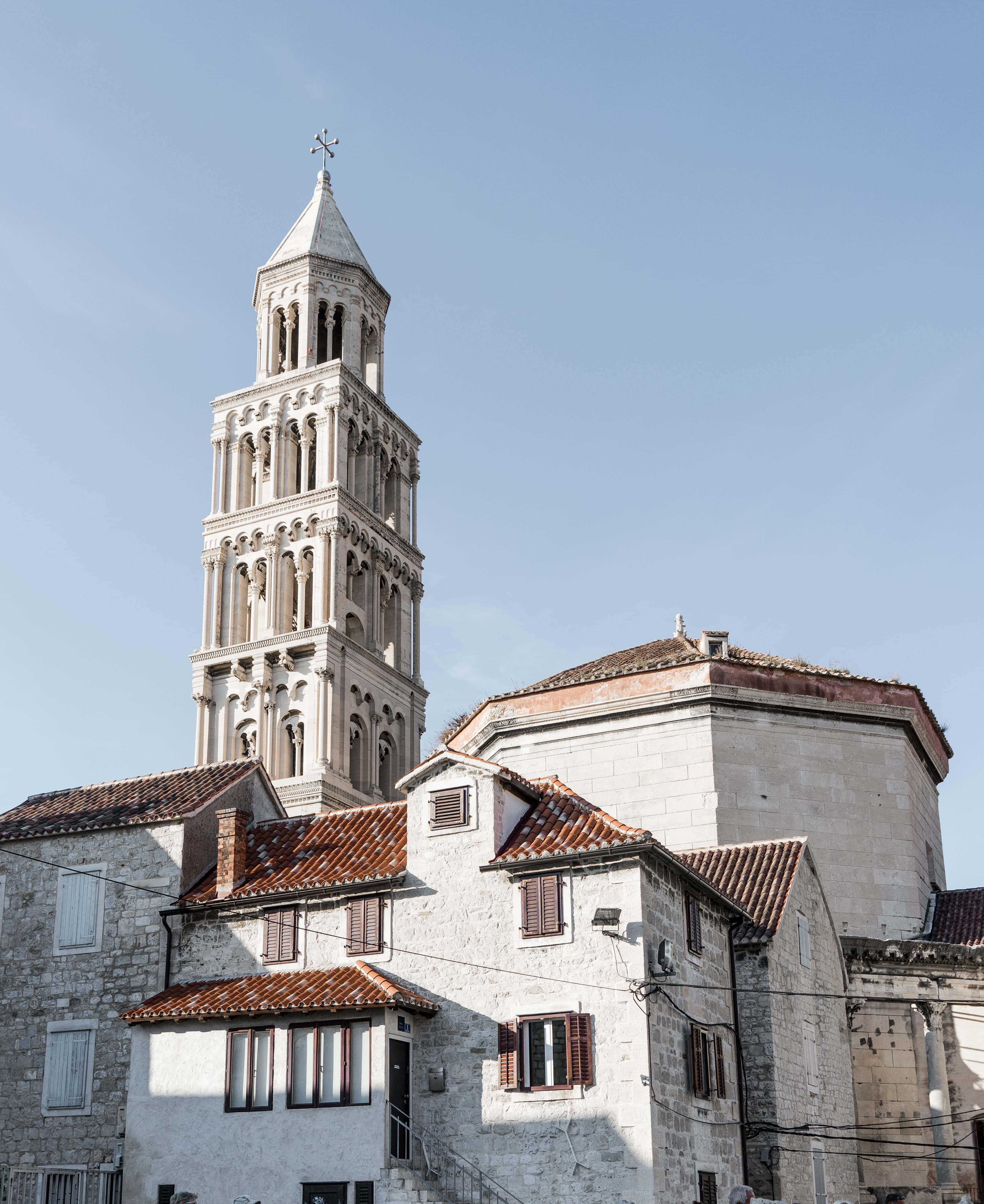 Croatia 桌布 壁紙 — 一座高聳的石造鐘樓，帶有拱形窗戶，矗立在白色粉刷的建築物之上，這些建築物擁有陶土屋頂，背景是清澈的藍天。