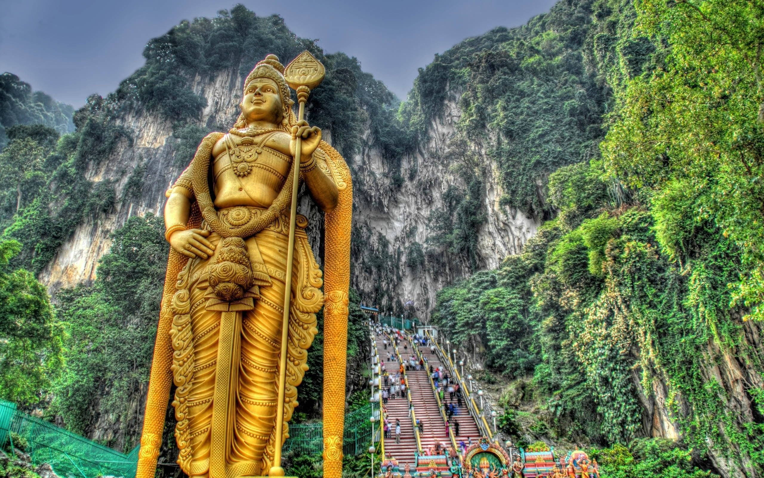 Tamil God 桌布 壁紙 — 一座高聳的穆魯甘勳爵金色雕像雄偉地矗立在鬱鬱蔥蔥的綠色山腳下，遊客正在攀登樓梯。