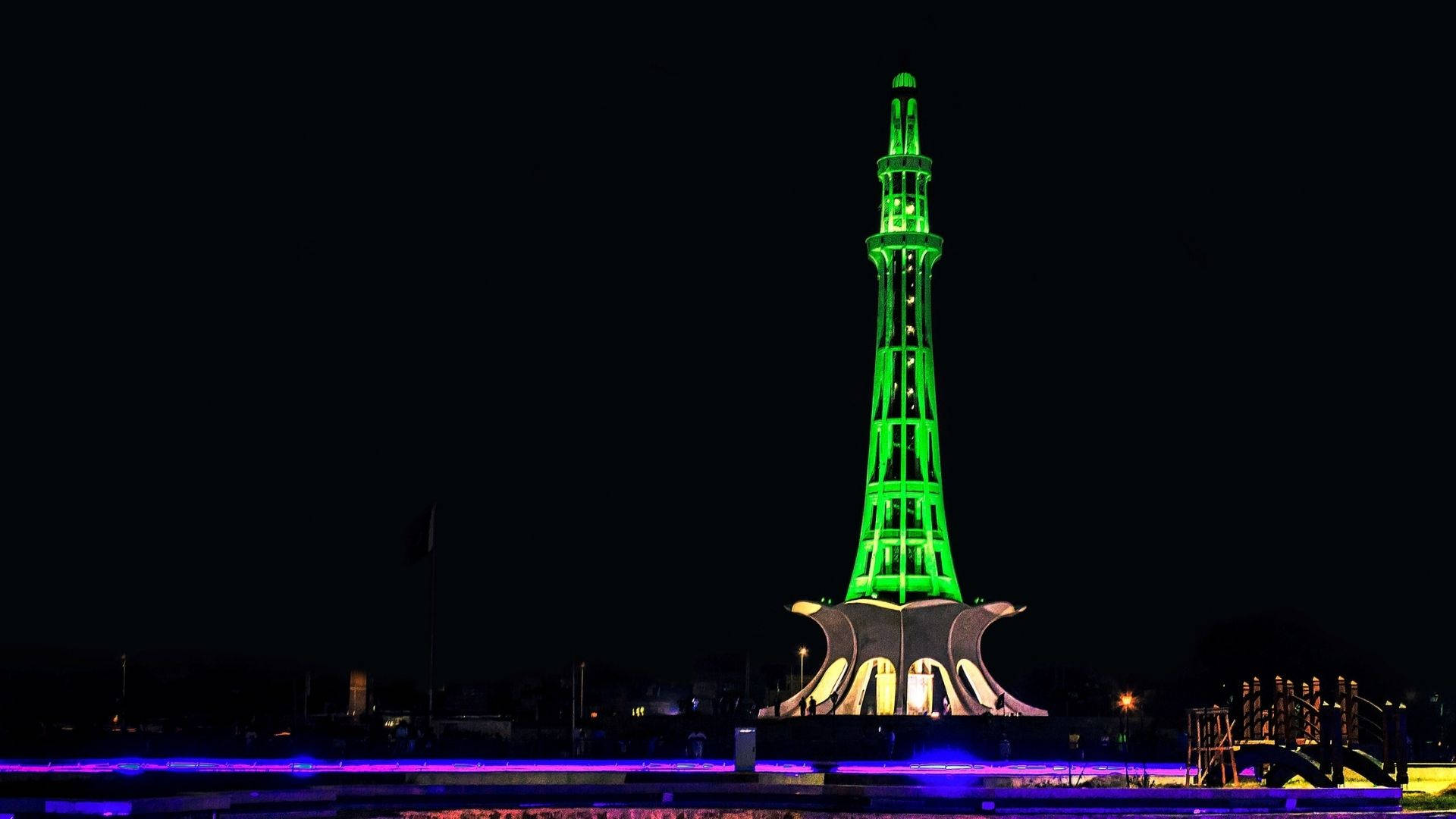 Pakistan 桌布 壁紙 — 一座高聳的紀念碑在漆黑的夜空中閃耀著鮮綠色，紫色燈光沿著其底部延伸。