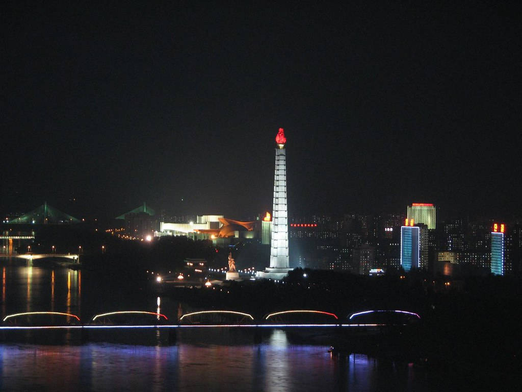 韓國 桌布 壁紙 — 一座高聳的紀念碑頂端有紅色火焰，在夜空中閃耀，倒映在下方水面，背景是燈火通明的橋樑和城市景觀。