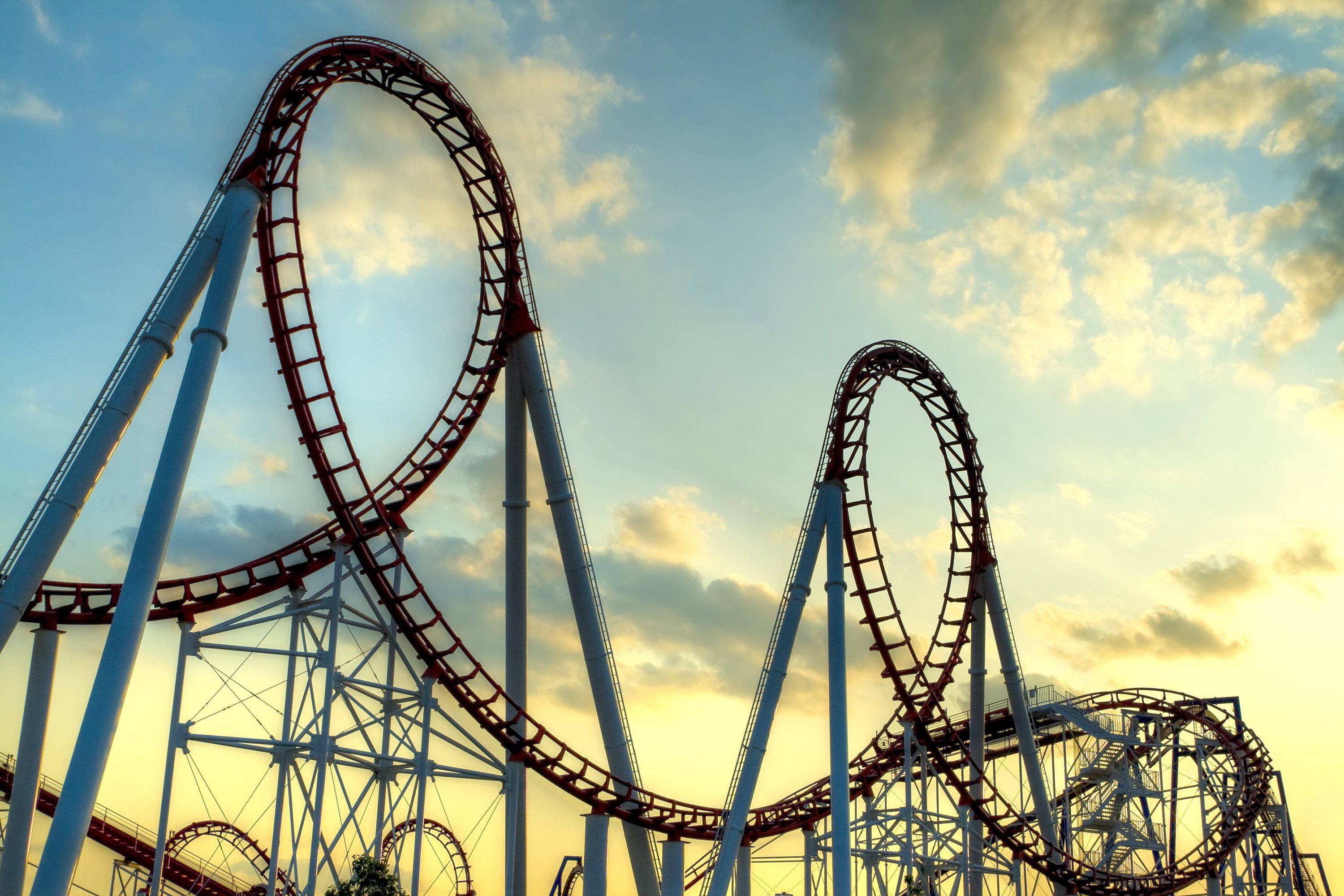 Roller Coaster 桌布 壁紙 — 一座高聳的紅白相間雲霄飛車在金色日落天空和散落的雲朵中蜿蜒盤旋。