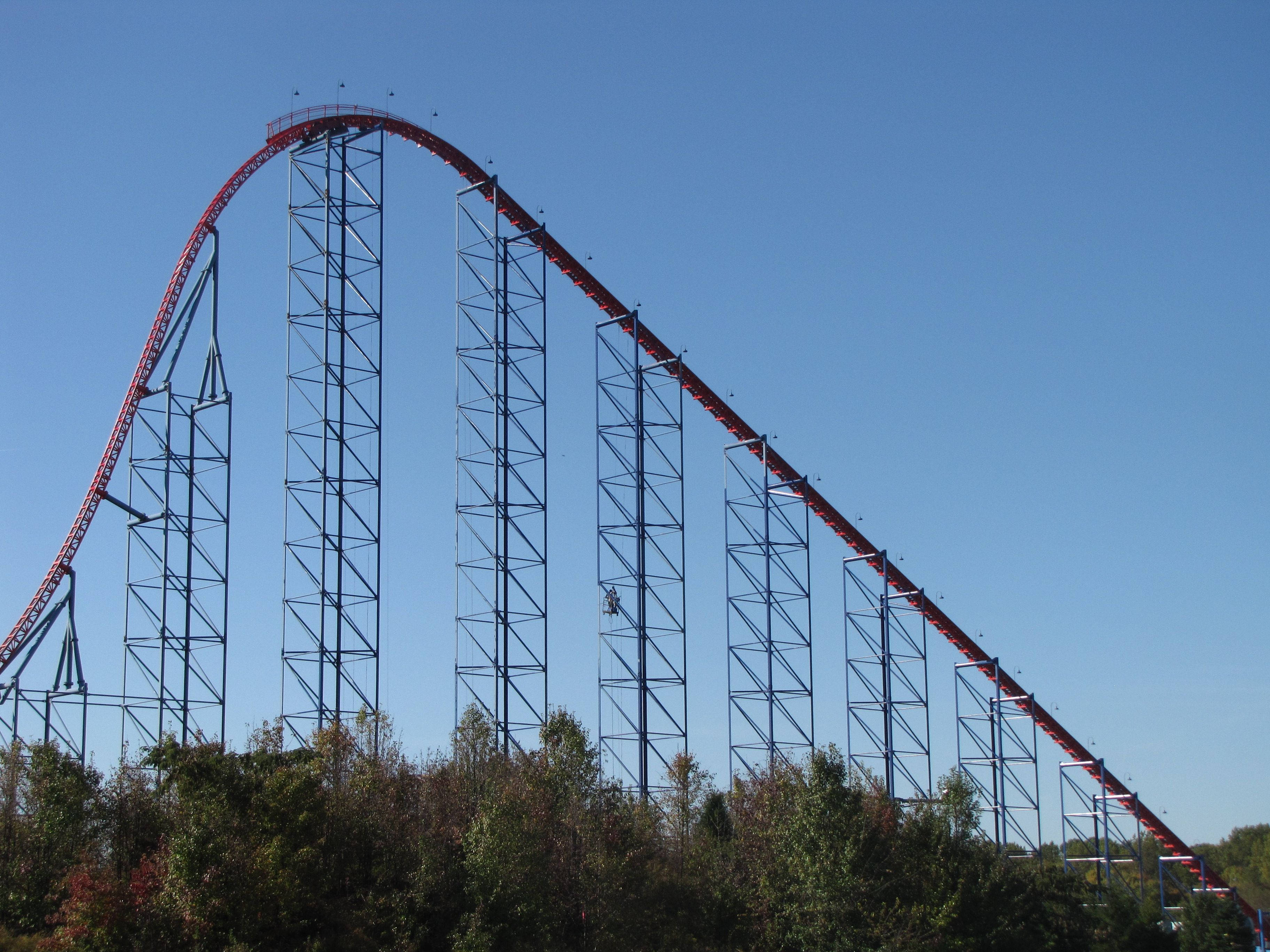Roller Coaster 桌布 壁紙 — 一座高聳的紅色雲霄飛車，帶有鋼鐵支架，在秋色樹木上方清澈的藍天下戲劇性地拱起。