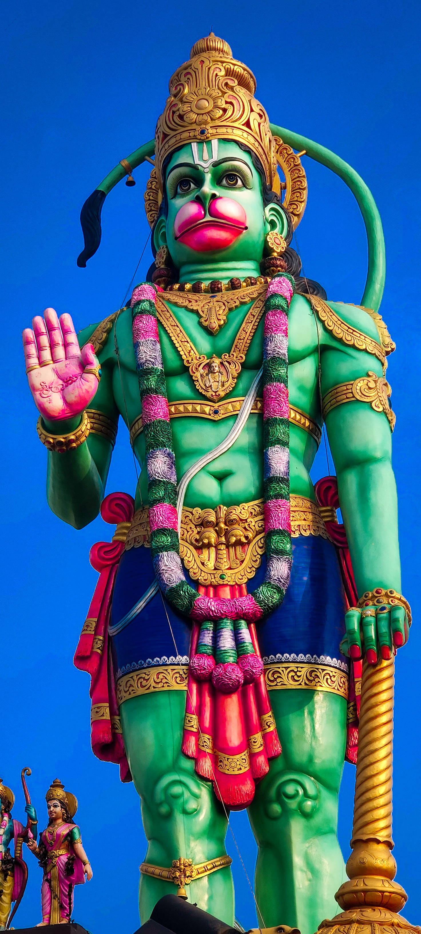 hanuman ji HD 桌布 壁紙 — 一座高聳的綠色哈努曼雕像，飾有金色飾物和粉紅色嘴唇，矗立在清澈的藍天下。