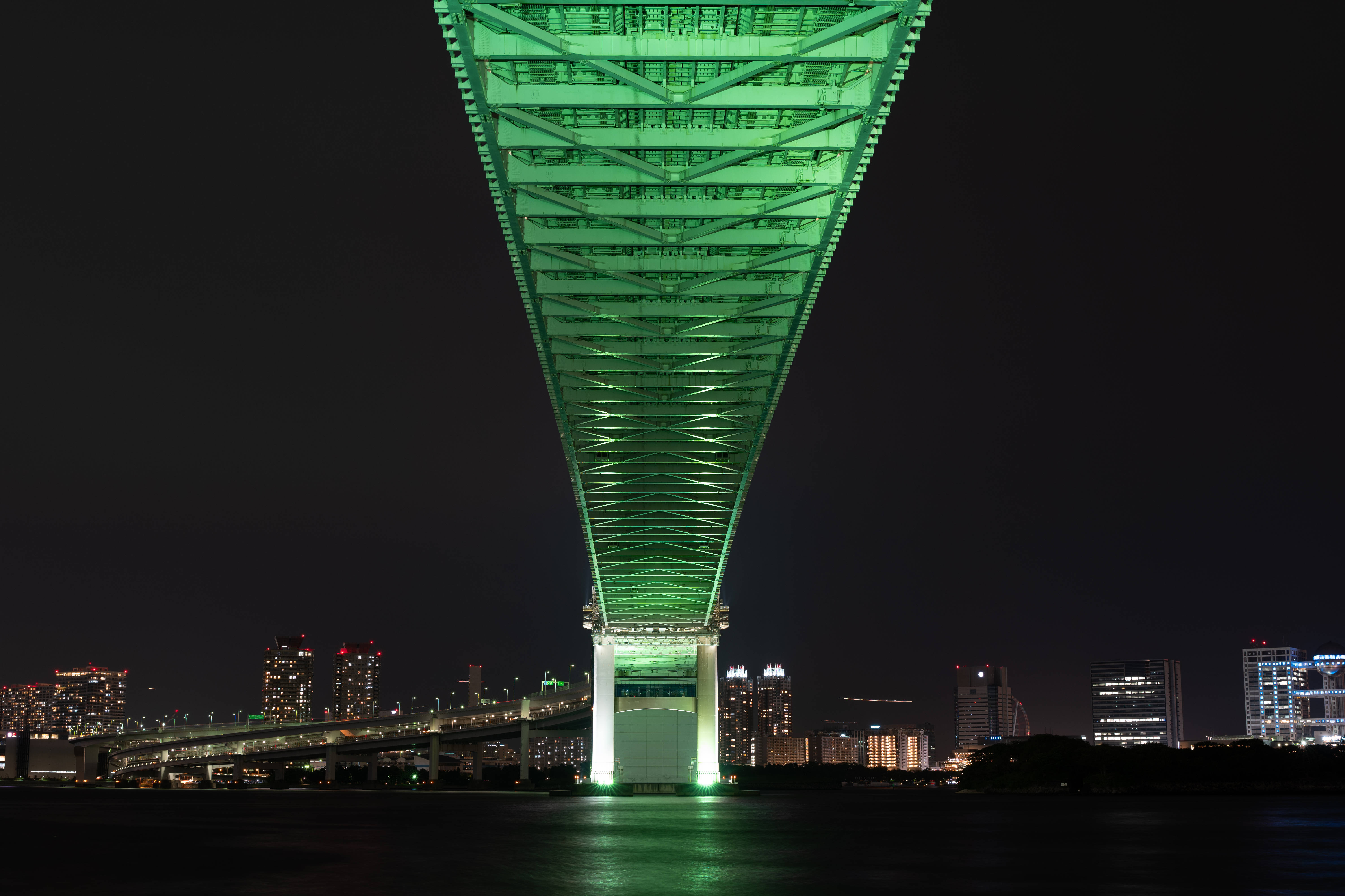 日本 桌布 壁紙 — 一座高聳的綠色照明橋樑在夜間拱跨水面，背景是閃爍的城市天際線。