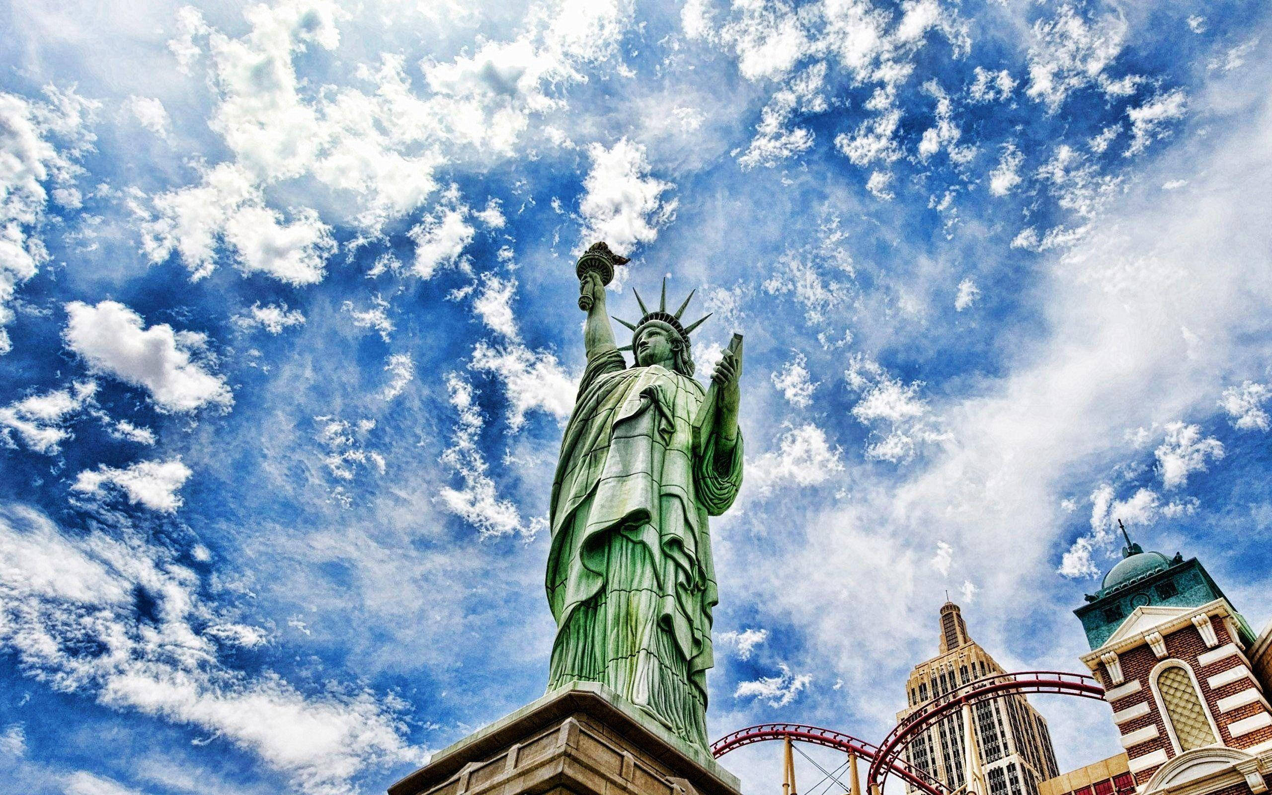 New York 桌布 壁紙 — 一座高聳的綠色自由女神像，在充滿蓬鬆白雲的鮮豔藍天下傲然矗立。