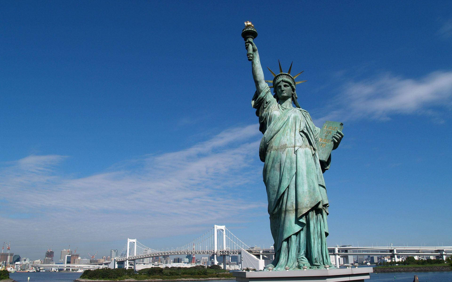 New York 桌布 壁紙 — 一座高聳的綠色自由女神像在晴朗的藍天下傲然矗立，背景是現代化的懸索橋和城市天際線。