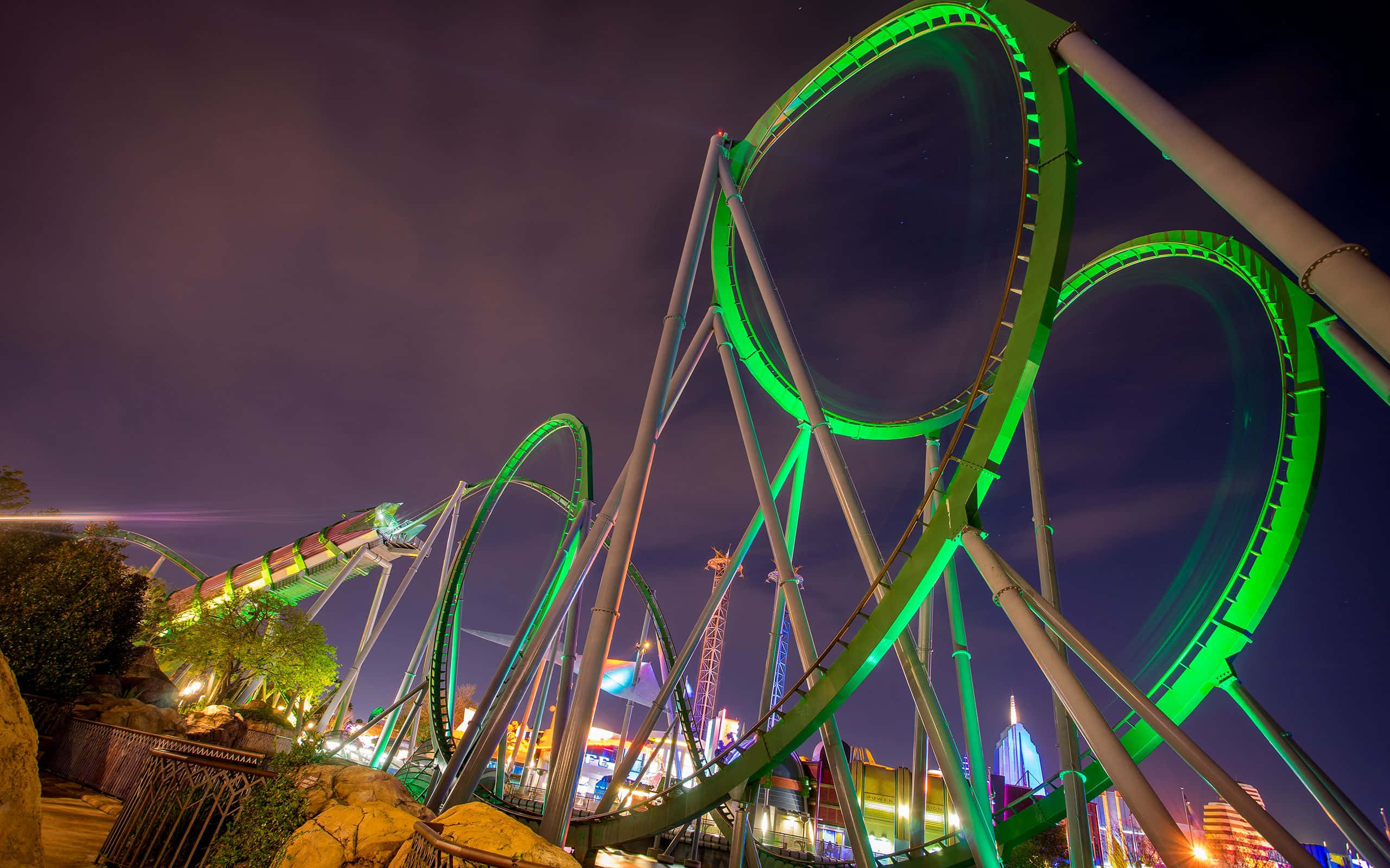 Roller Coaster 桌布 壁紙 — 一座高聳的綠色雲霄飛車在夜燈下發光，背景是黑暗的天空和遠處的城市建築。
