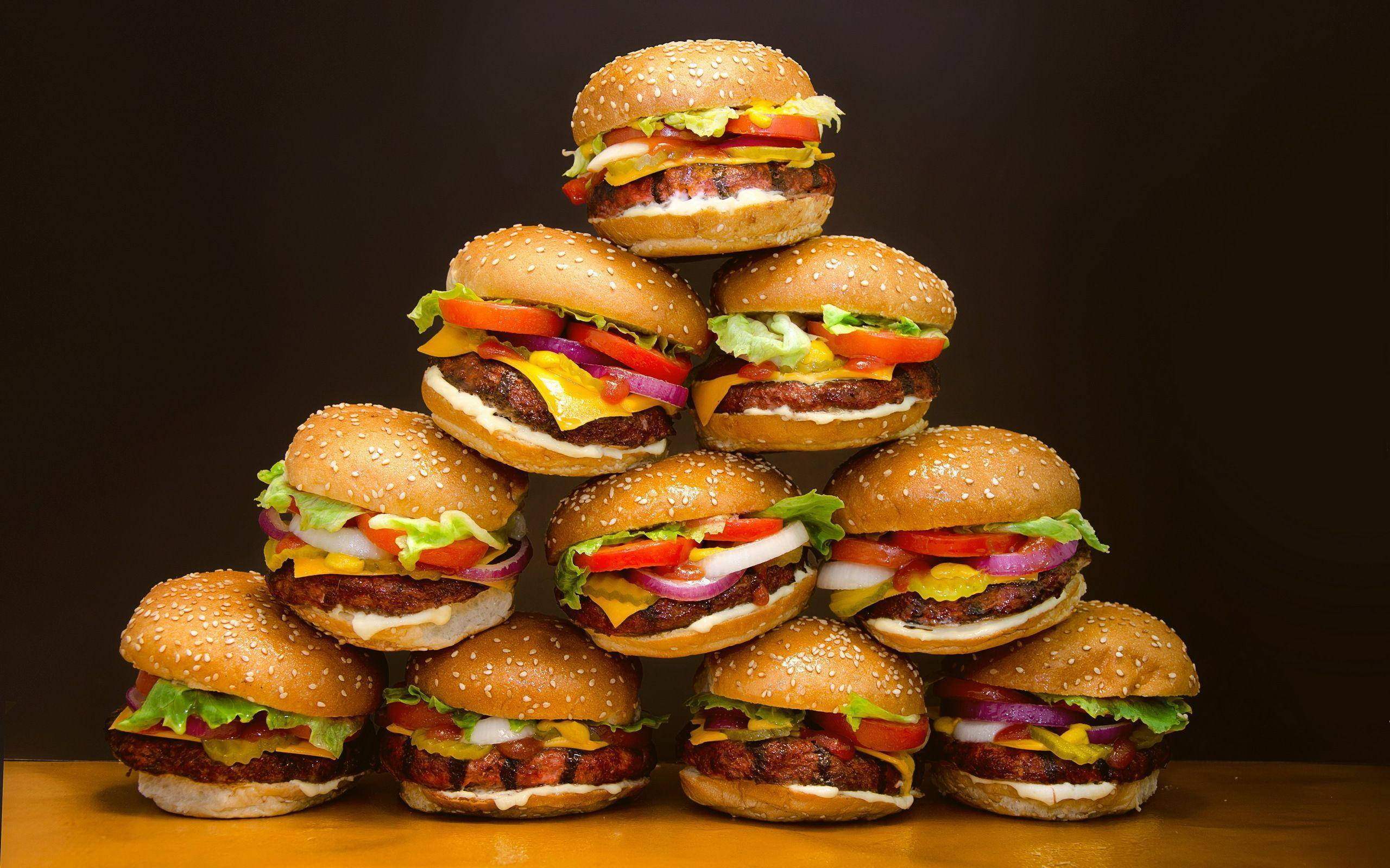 Burger 桌布 壁紙 — 一座高聳的芝麻漢堡金字塔，堆滿生菜、番茄、起司和洋蔥，襯托在深色背景前。