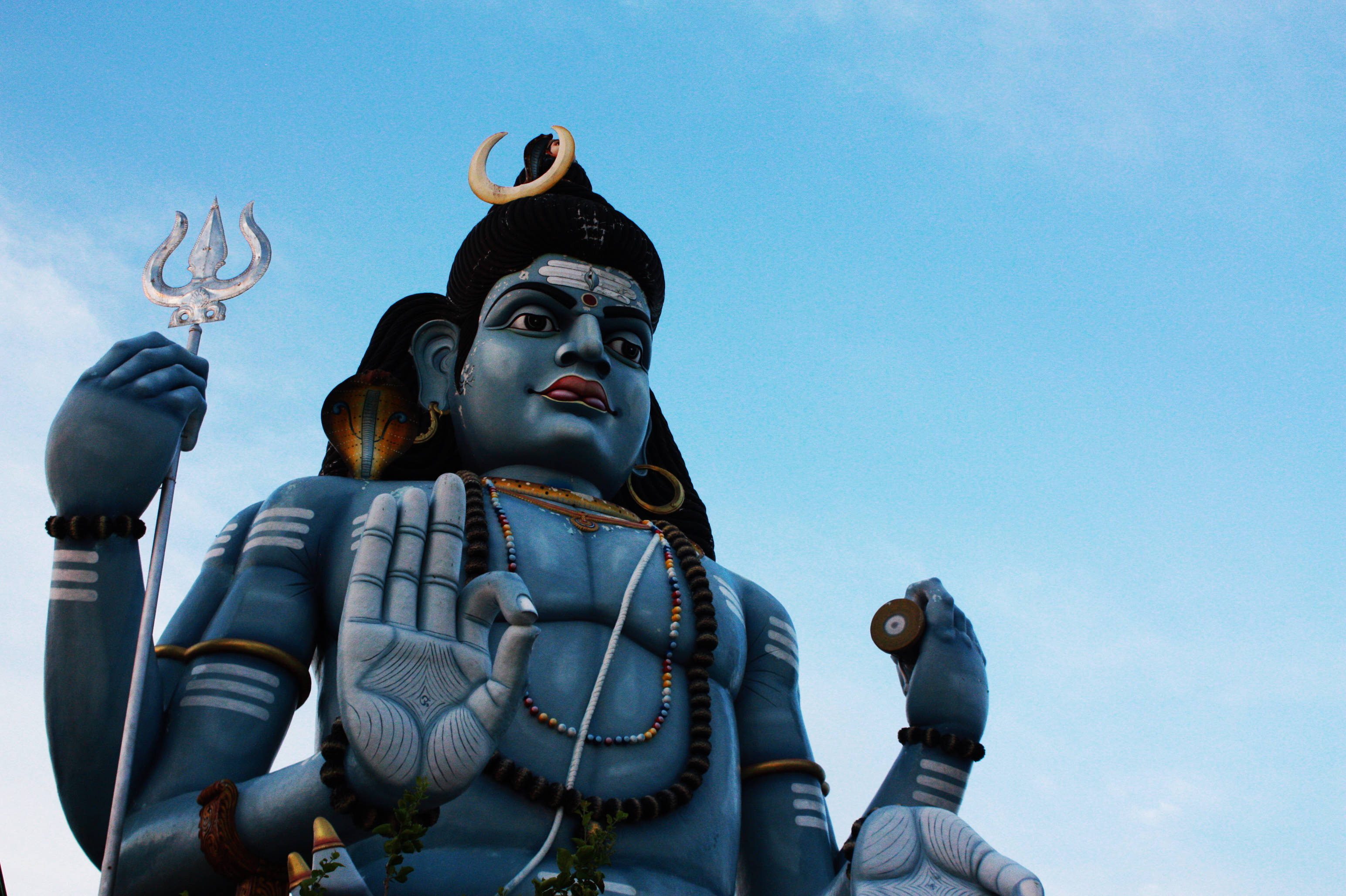 adiyogi shiva 桌布 壁紙 — 一座高聳的藍色濕婆神像，手持三叉戟、新月，舉起手，背景是清澈的藍天。