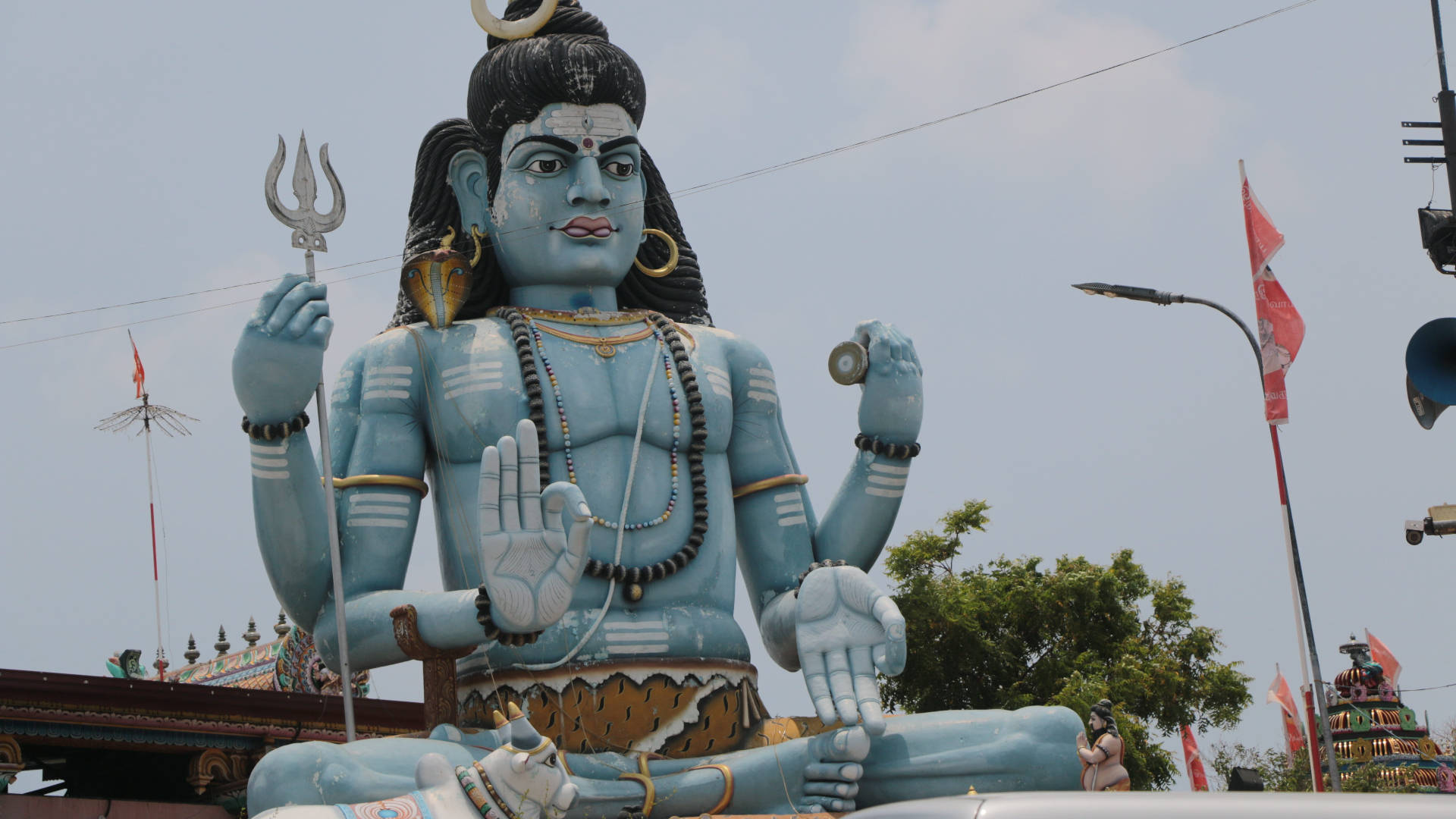 Hindu Temple 桌布 壁紙 — 一座高聳的藍色濕婆神像，手持三叉戟和鼓，寧靜地坐在明亮的天空下，附近的寺廟旗幟隨風飄揚。