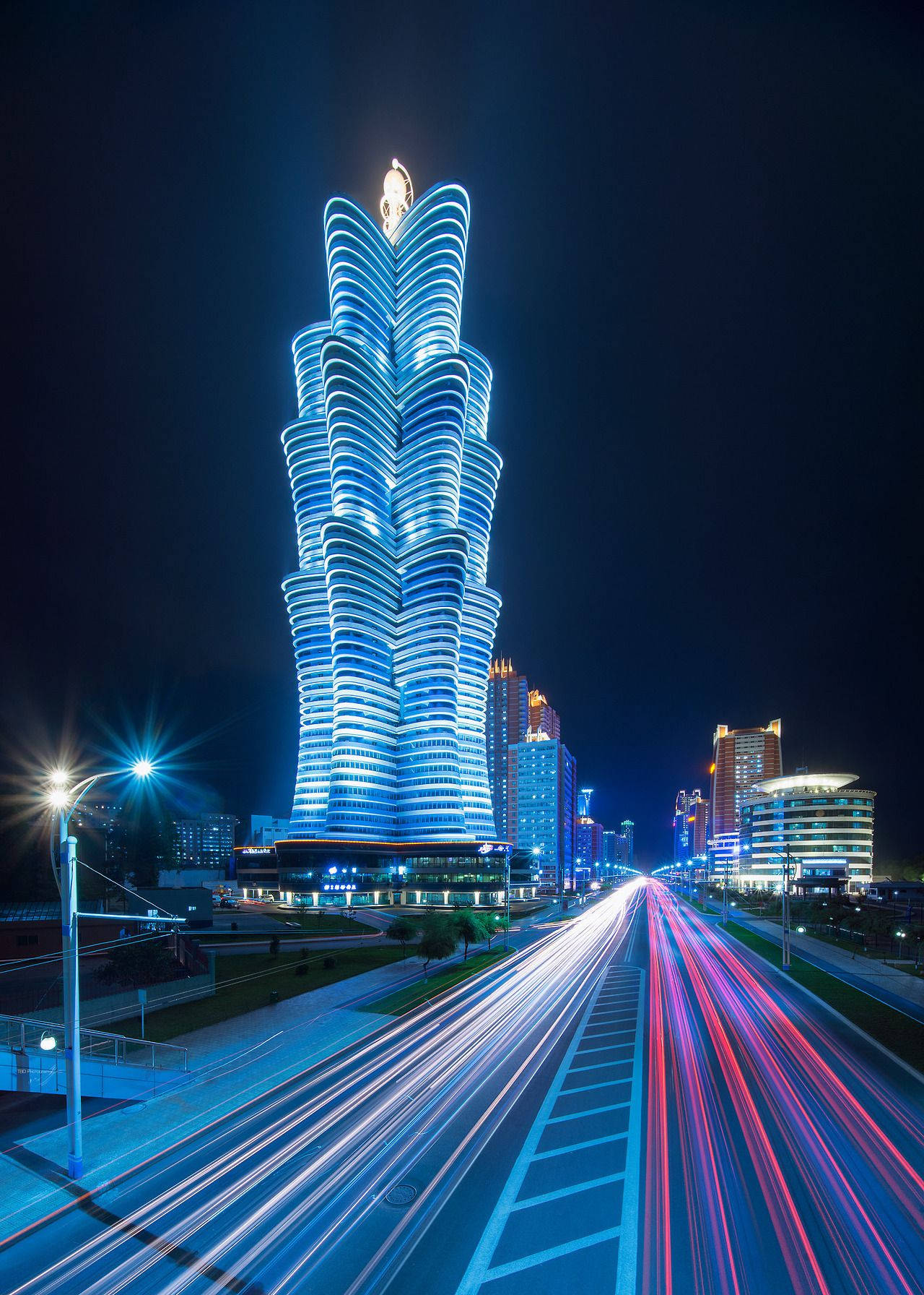韓國 桌布 壁紙 — 一座高聳的藍色發光摩天大樓主導著夜景，下方車流留下紅色和白色的光軌。