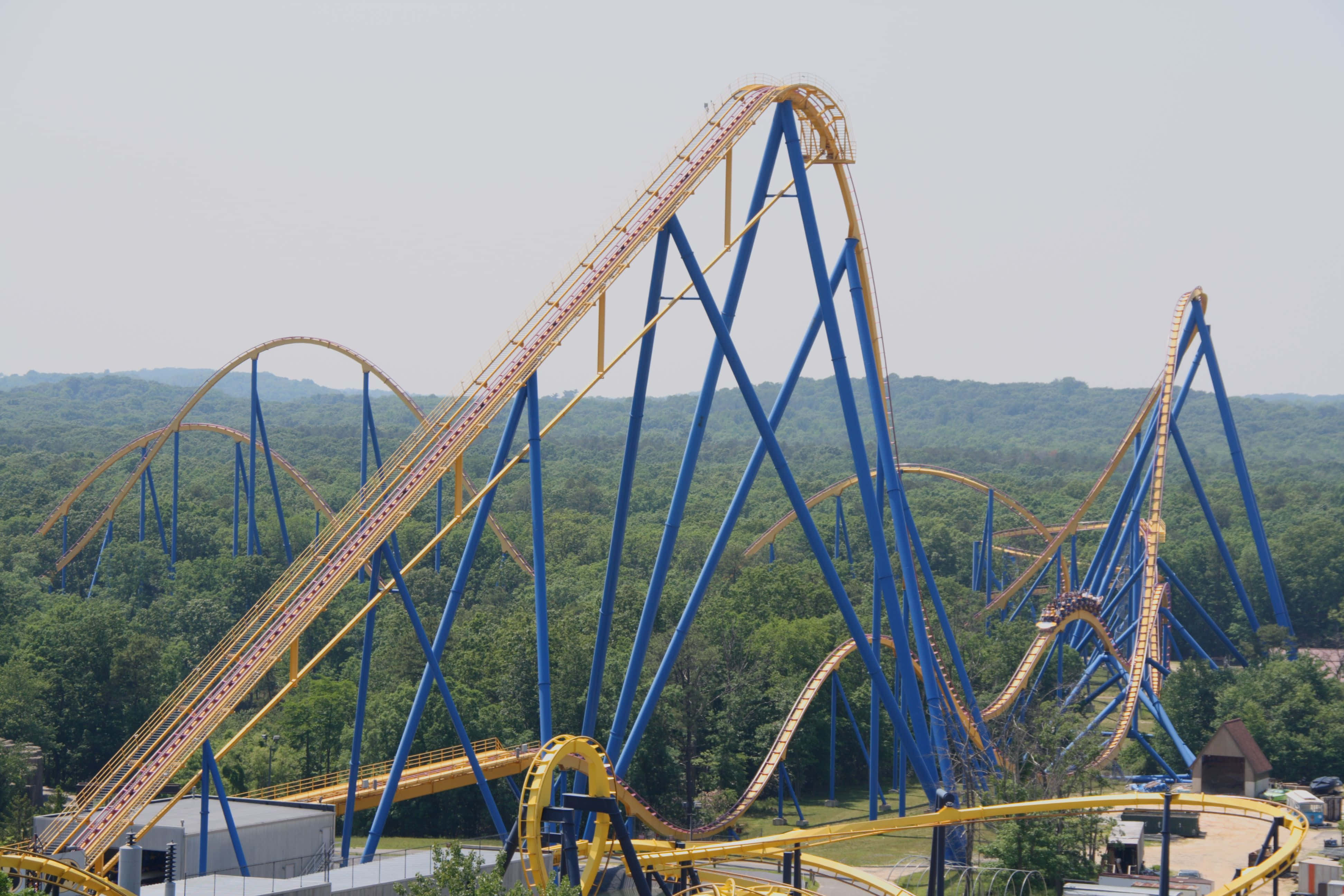 Six Flags 桌布 壁紙 — 一座高聳的藍黃色雲霄飛車在朦朧的天空下，以茂密的綠色山丘為背景，扭轉和環繞。
