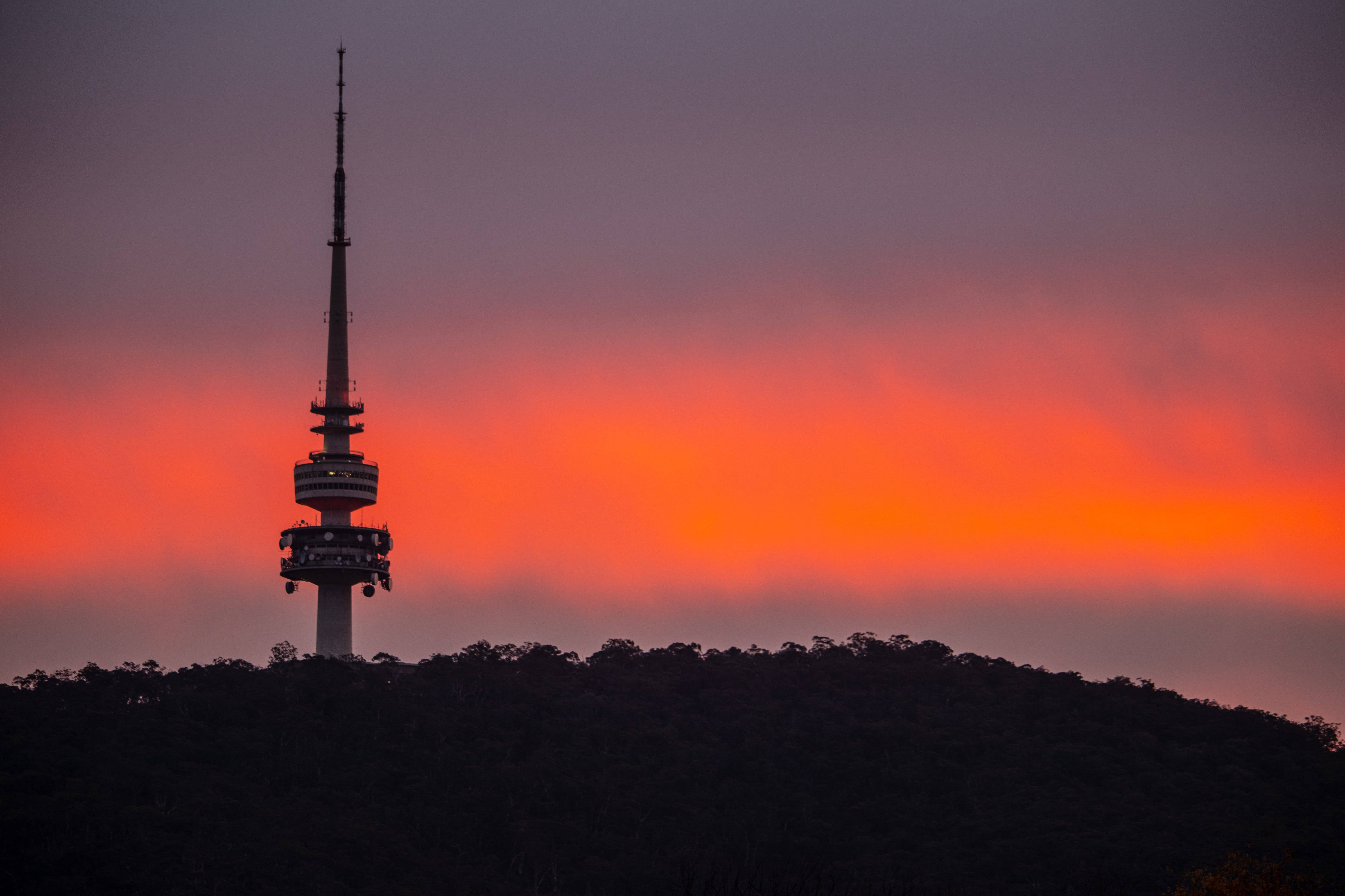 Canberra 桌布 壁紙 — 一座高聳的通訊塔在黑暗森林覆蓋的山丘上，以火熱的橙色和紫色日落天空為背景，呈現剪影。
