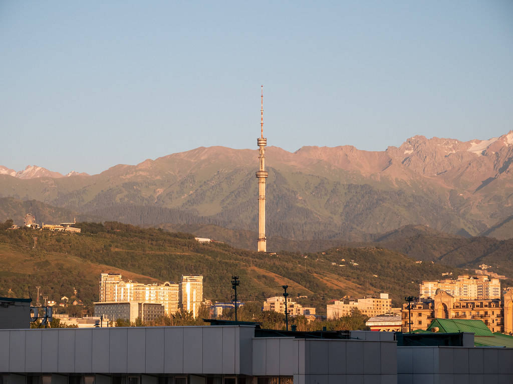Kazakhstan 桌布 壁紙 — 一座高聳的通訊塔矗立於城市景觀之上，現代建築依偎在崎嶇山脈的背景下，沐浴在金色的日落光芒中。