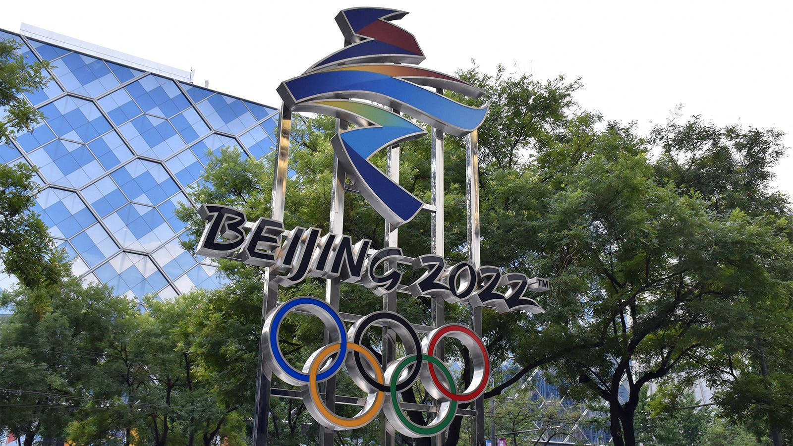 Winter Olympics 桌布 壁紙 — 一座高聳的金屬北京2022奧運標誌雕塑，帶有環環相扣的奧運五環，矗立在茂密的綠樹叢中，旁邊是一座現代化的藍色玻璃建築。