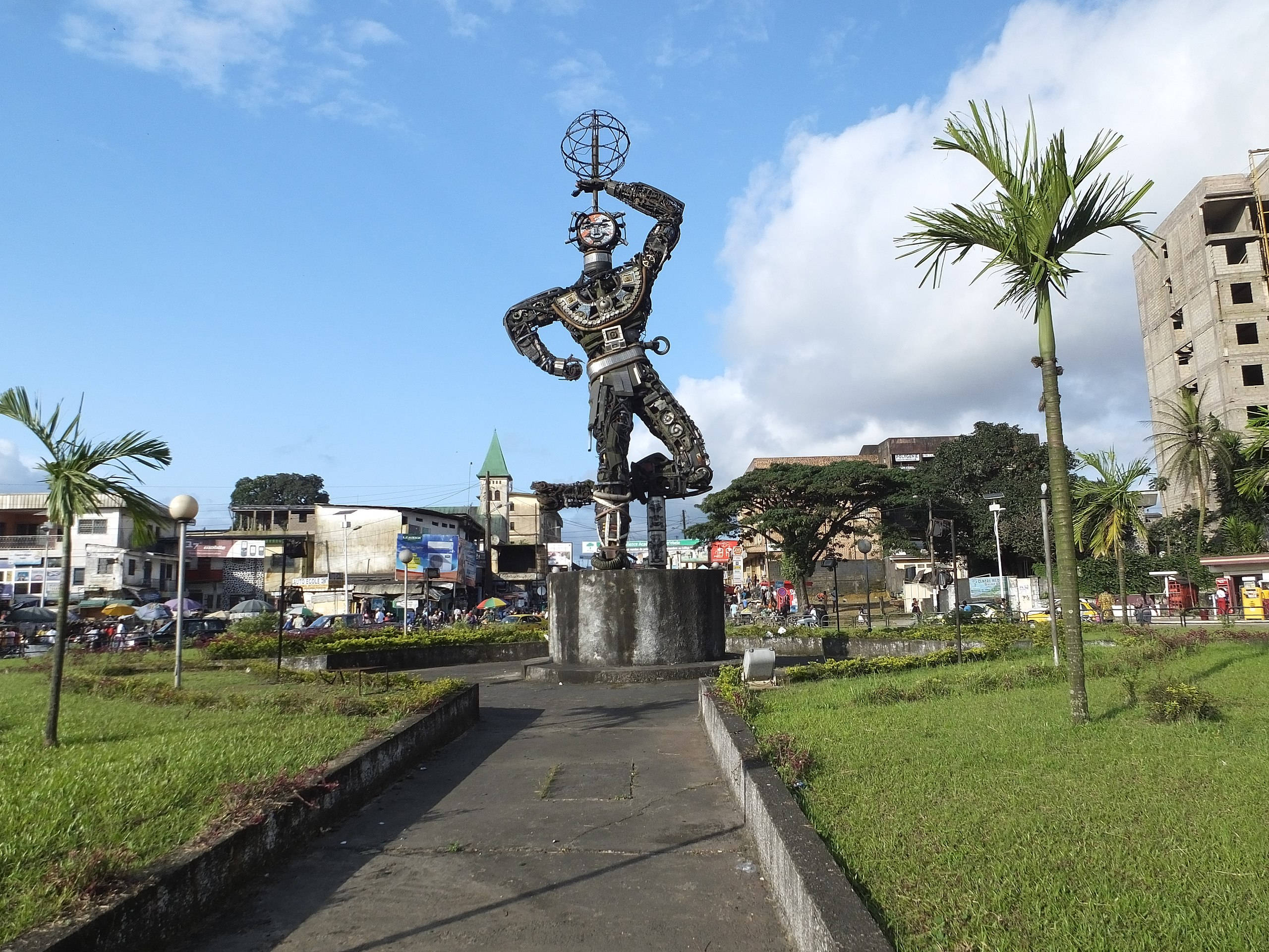 Statue Aesthetic 桌布 壁紙 — 一座高聳的金屬雕像，人物頭頂平衡著一個地球儀，矗立在綠草如茵的城市廣場上，天空湛藍，白雲點點。