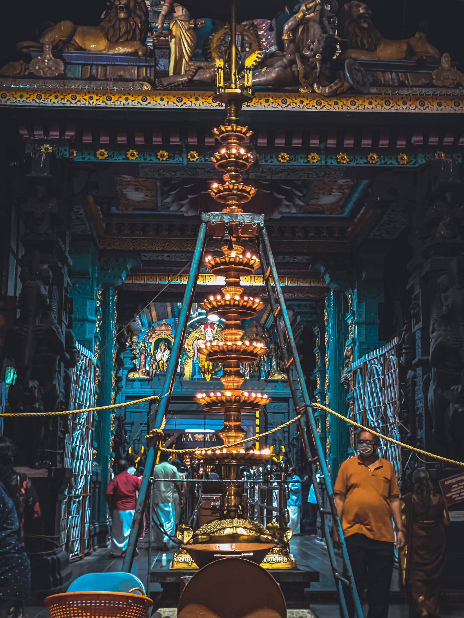 Hindu Temple 桌布 壁紙 — 一座高聳的金色儀式燈在雕刻精美的寺廟內溫暖地發光，寺廟有深藍色的牆壁，附近有朝拜者。