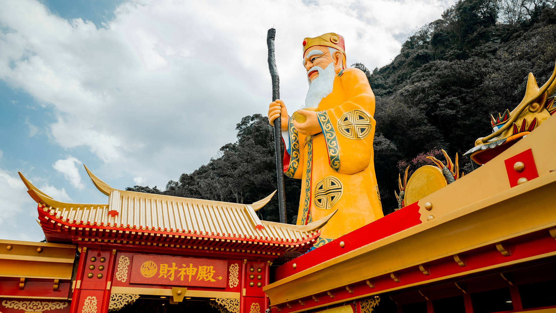 金色神祇雕像手持權杖，位於裝飾華麗的紅色寺廟頂上，背景是多雲的天空。