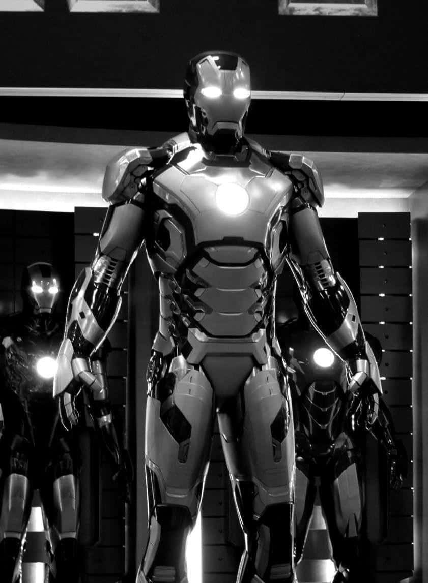 Iron Man Suit 桌布 壁紙 — 一座高聳的鋼鐵人裝甲在單色燈光下矗立，兩側還有另外兩套裝甲，位於高科技艙室中。