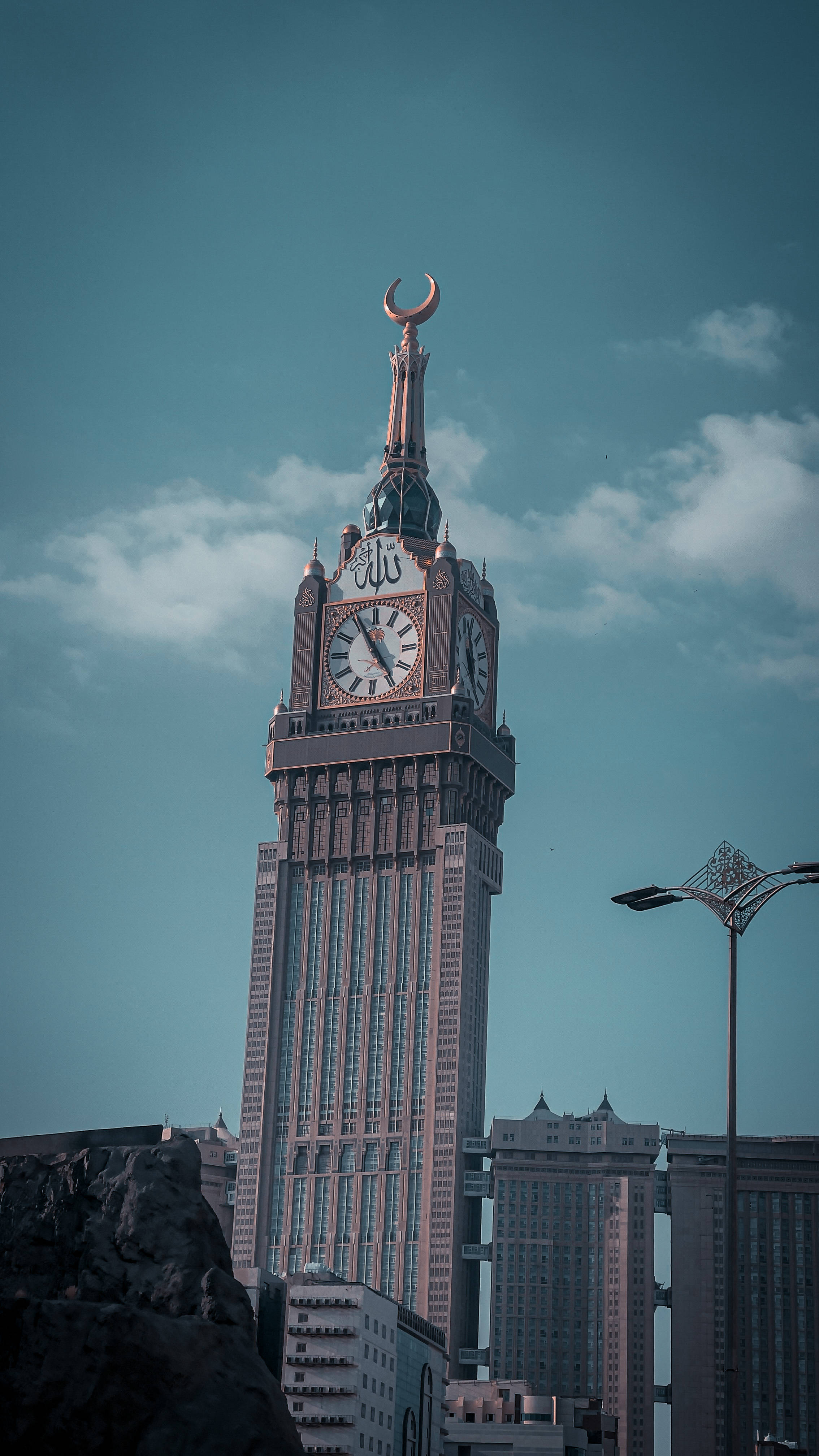Makkah 桌布 壁紙 — 一座高聳的鐘樓，頂部有新月形尖頂，在帶有散雲的藍綠色天空下矗立。