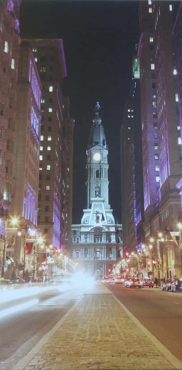 Philly 桌布 壁紙 — 一座高聳的鐘樓在夜間被照亮，矗立在高樓大廈之間，鵝卵石街道上行駛的汽車留下光跡。