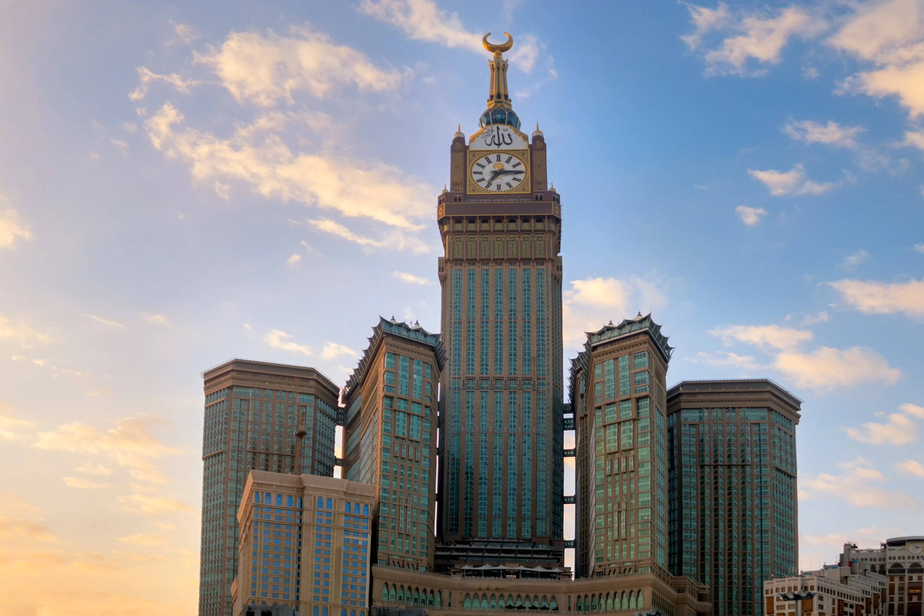 Saudi 桌布 壁紙 — 一座高聳的鐘樓帶有新月形頂飾，在金色夕陽天空的映襯下，聳立於玻璃摩天大樓之上。