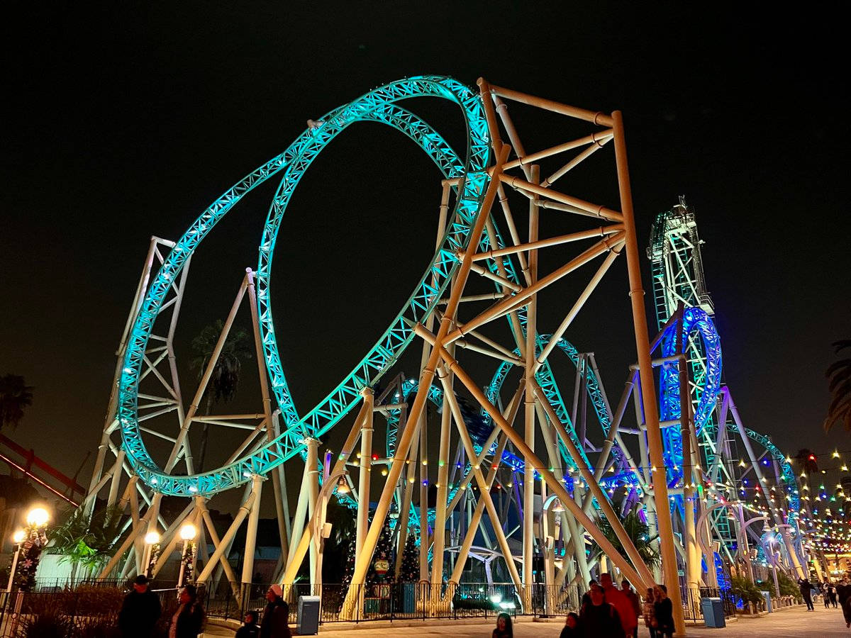 Roller Coaster 桌布 壁紙 — 一座高聳的雲霄飛車在充滿活力的青綠色和藍色燈光下照亮，主導了夜間遊樂園場景，遊客在下方行走。