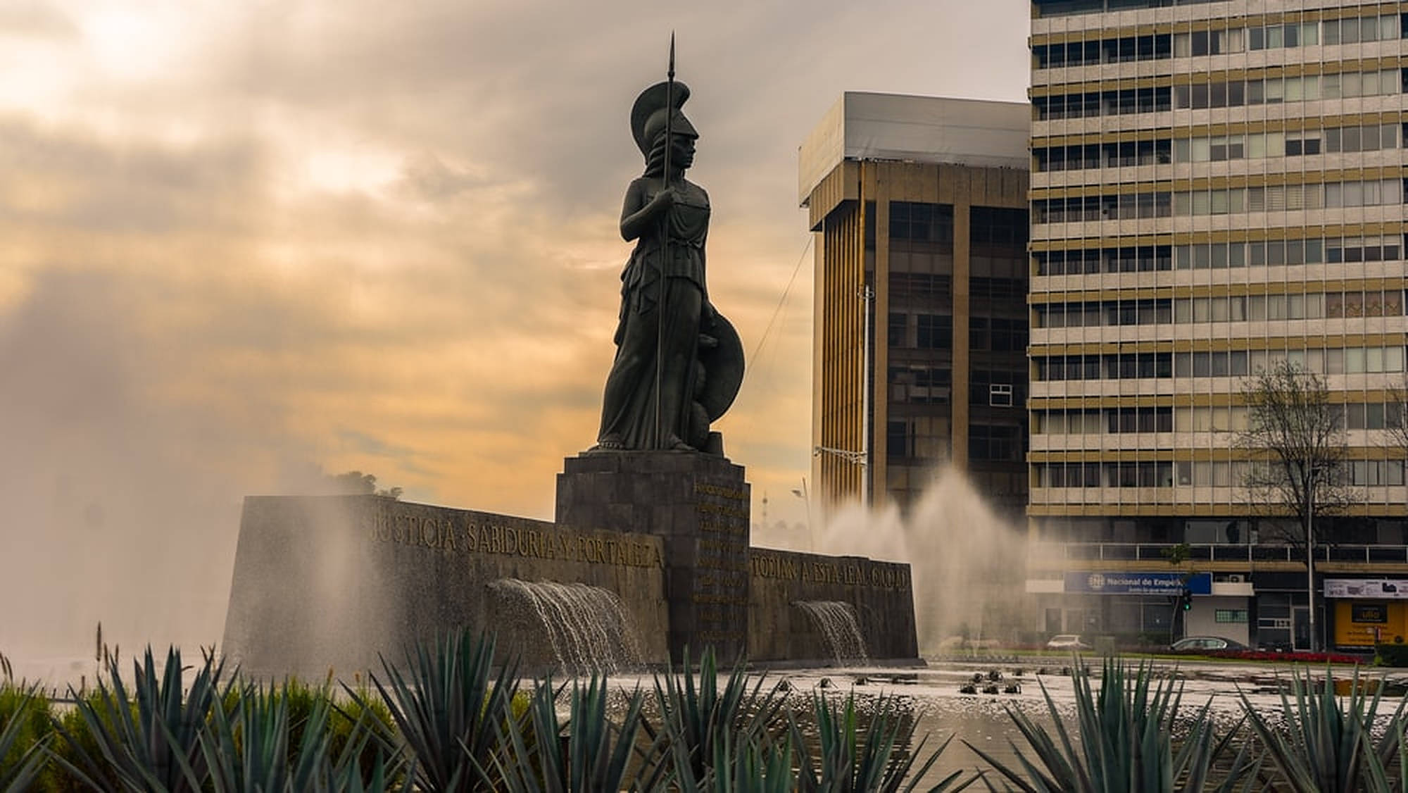 Guadalajara 桌布 壁紙 — 一座高聳的青銅雕像，人物手持長矛和盾牌，矗立在噴泉中，周圍是現代建築，天空多雲，夕陽西下。