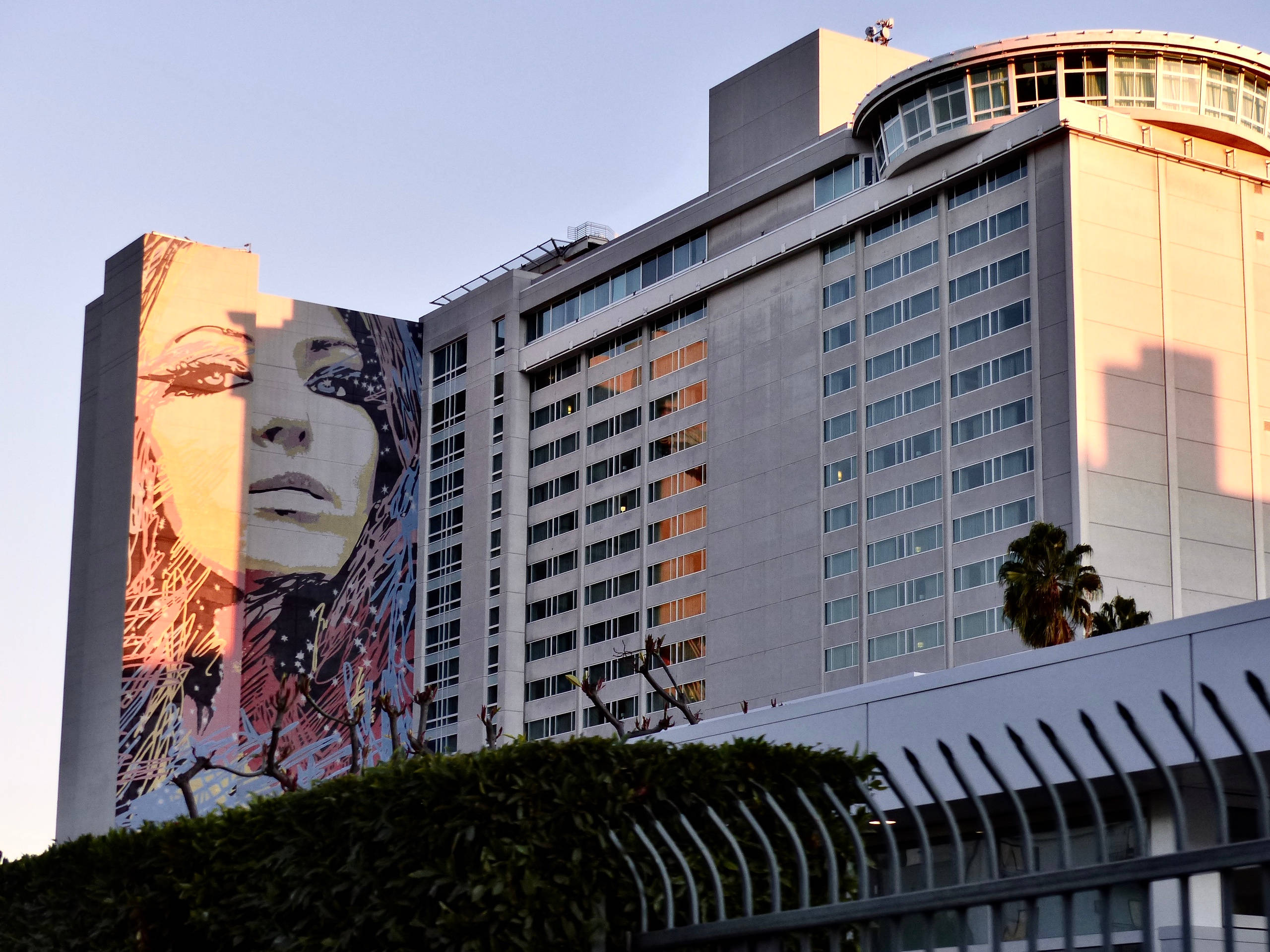 hollywood street 桌布 壁紙 — 一座高聳的飯店建築側面有一幅巨大的彩色女性面孔壁畫，沐浴在溫暖的日落光中。