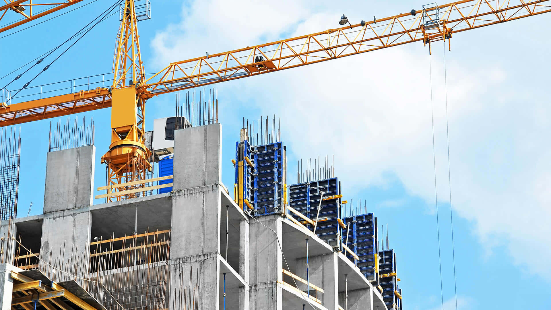 Building 桌布 壁紙 — 一座高聳的黃色起重機在明亮的藍天下，主導著一棟正在施工的多層混凝土建築，建築物有外露的鋼筋和藍色模板。