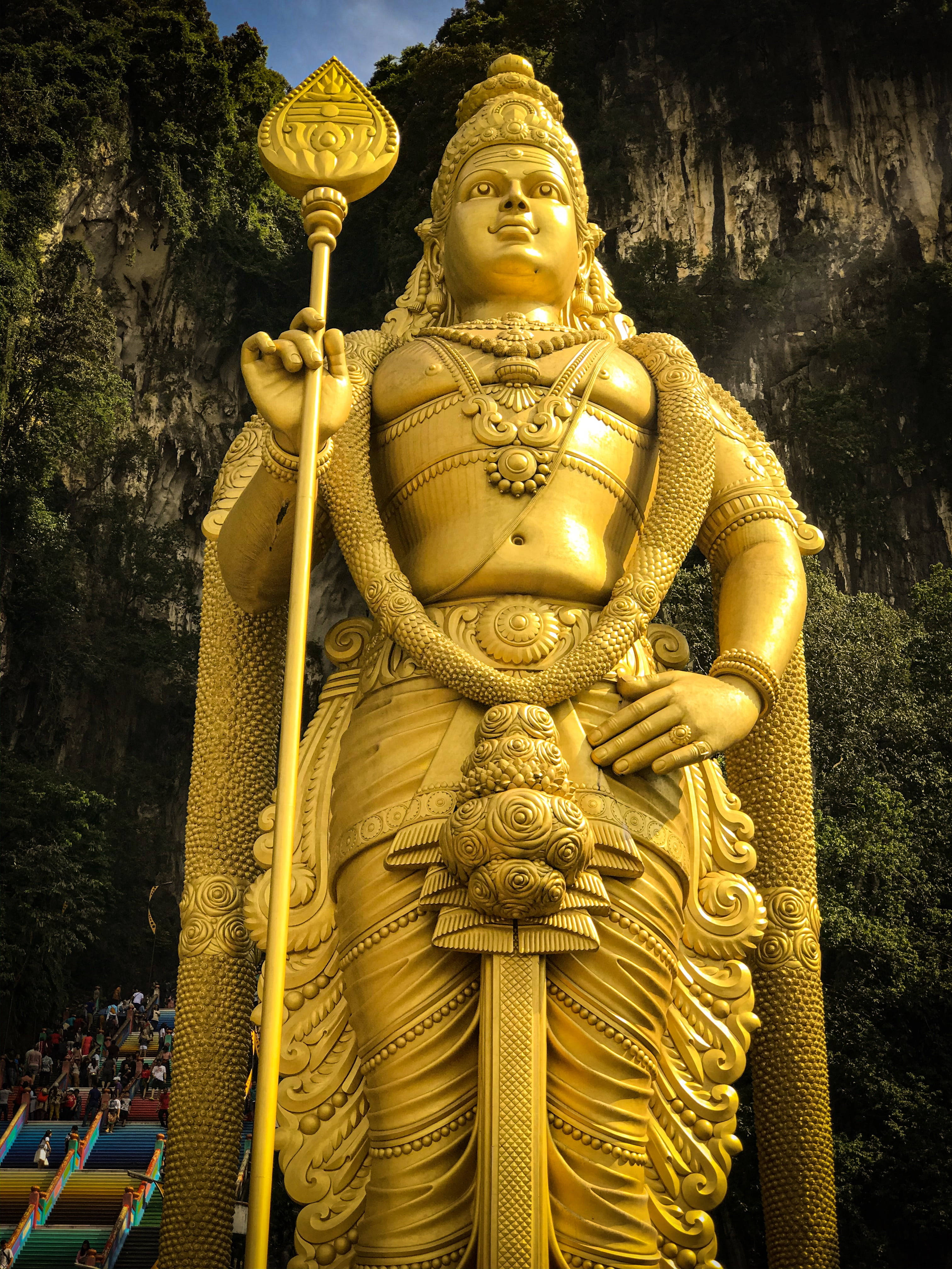 Tamil God 桌布 壁紙 — 一座高聳的黃金穆魯干勳爵雕像，雄偉地矗立在鬱鬱蔥蔥的綠色懸崖邊，下方有遊客攀爬色彩繽紛的階梯。