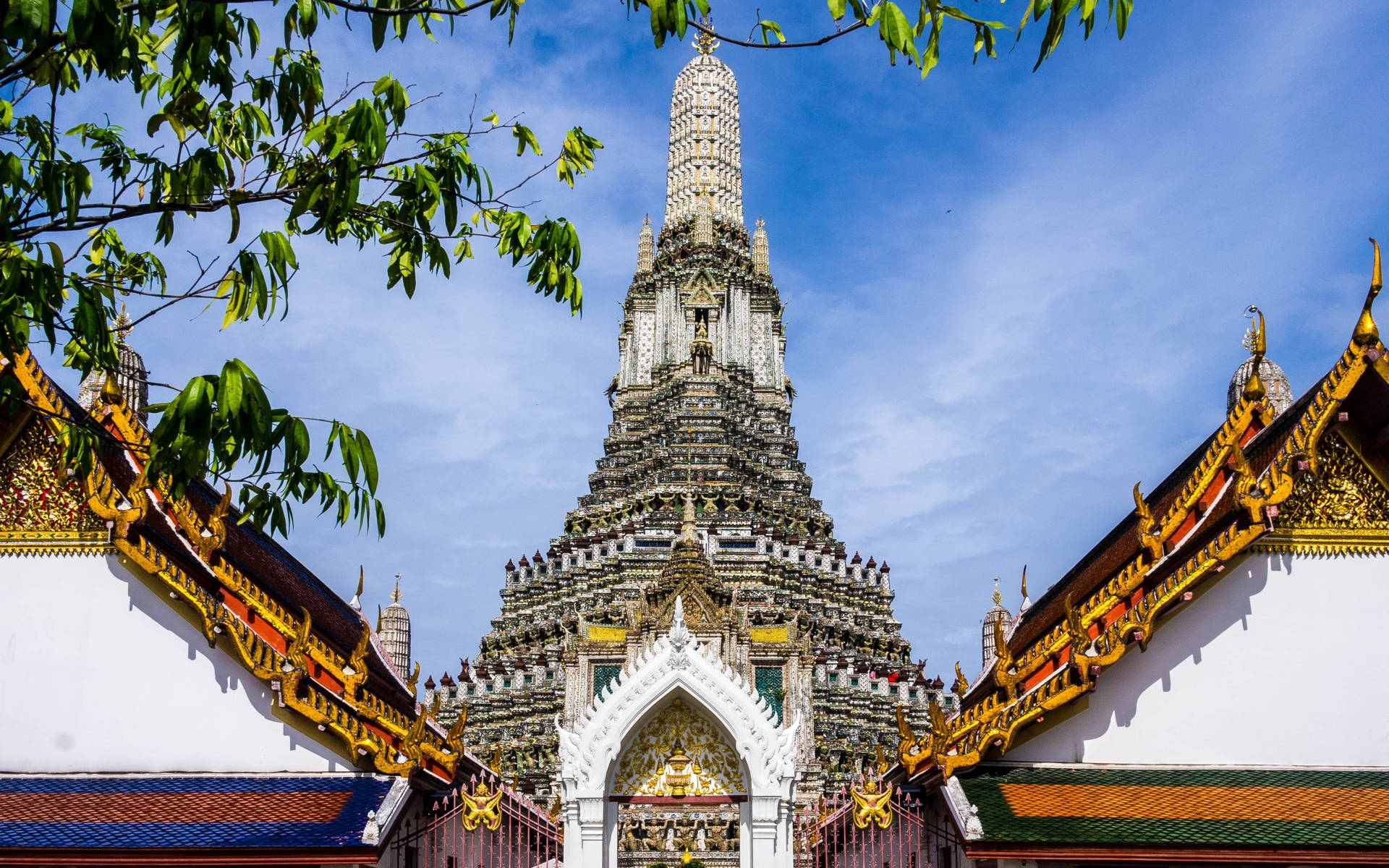 Bangkok 桌布 壁紙 — 一座高聳華麗的寺廟尖塔穿過茂密的綠葉，在金色屋頂結構框住的明亮藍天下矗立。
