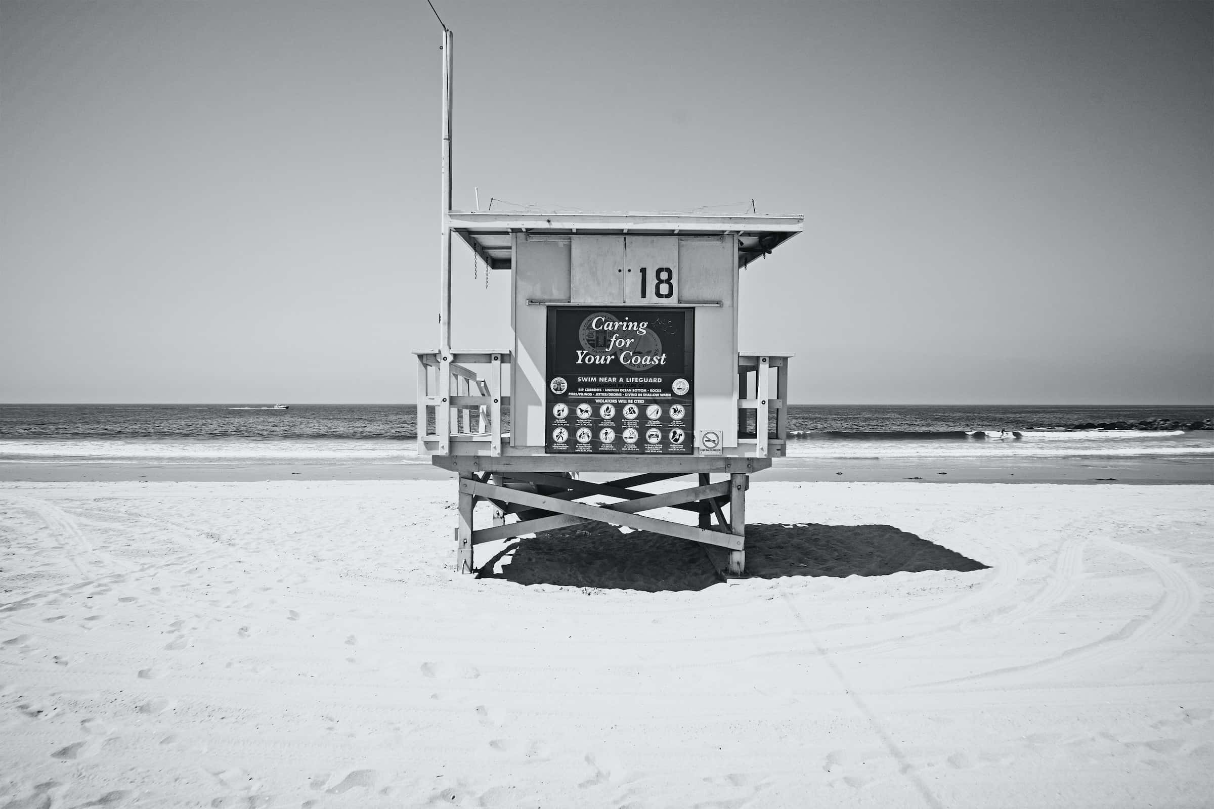 Black And White Beach 桌布 壁紙 — 一座鮮明的黑白救生塔獨自矗立在沙灘上，海浪在晴朗的天空下翻滾而來。