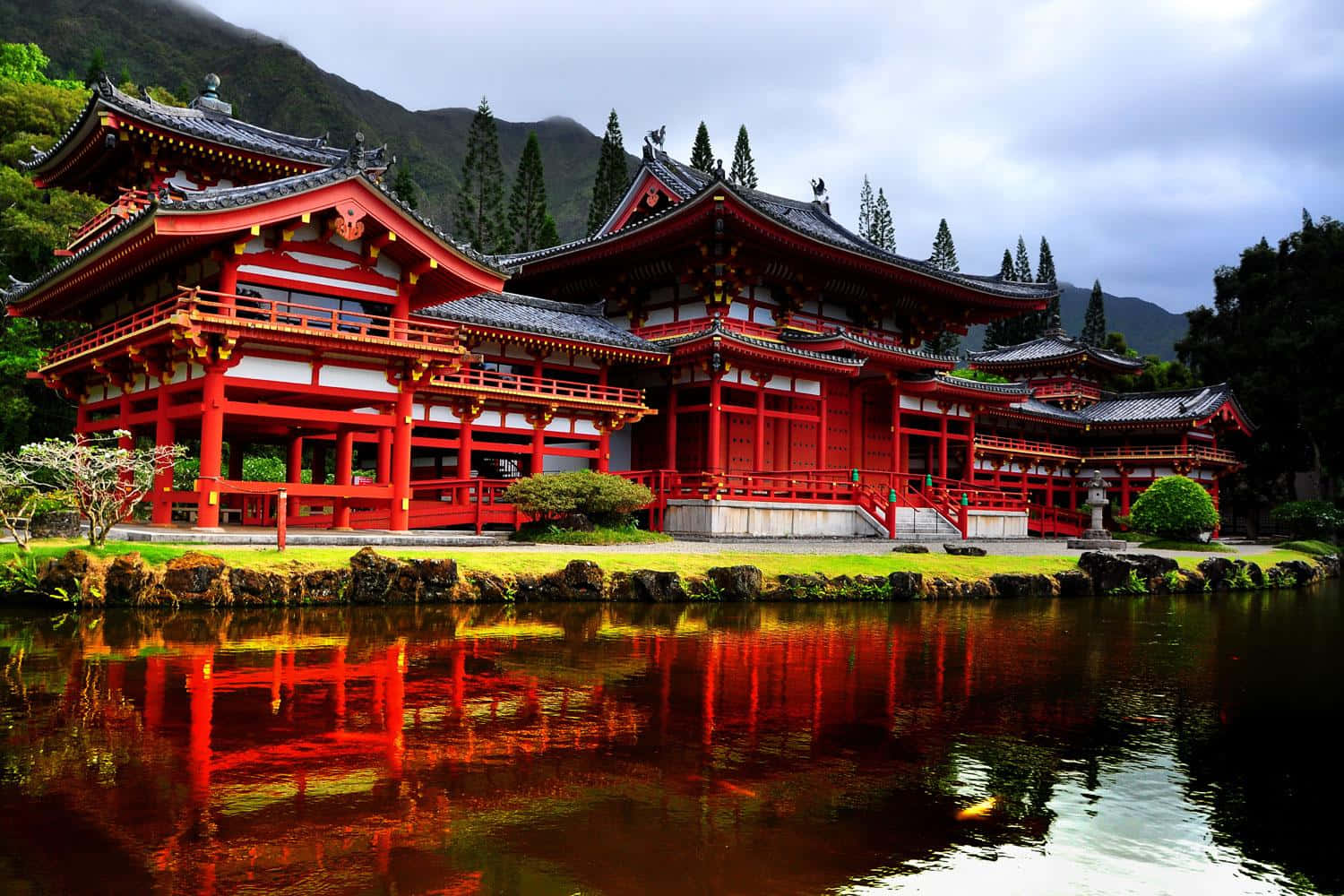 Feudal Japan 桌布 壁紙 — 一座鮮紅色的傳統日本寺廟，帶有黑色瓦片屋頂，完美倒映在平靜的池塘中，周圍環繞著綠植和霧氣繚繞的山脈。
