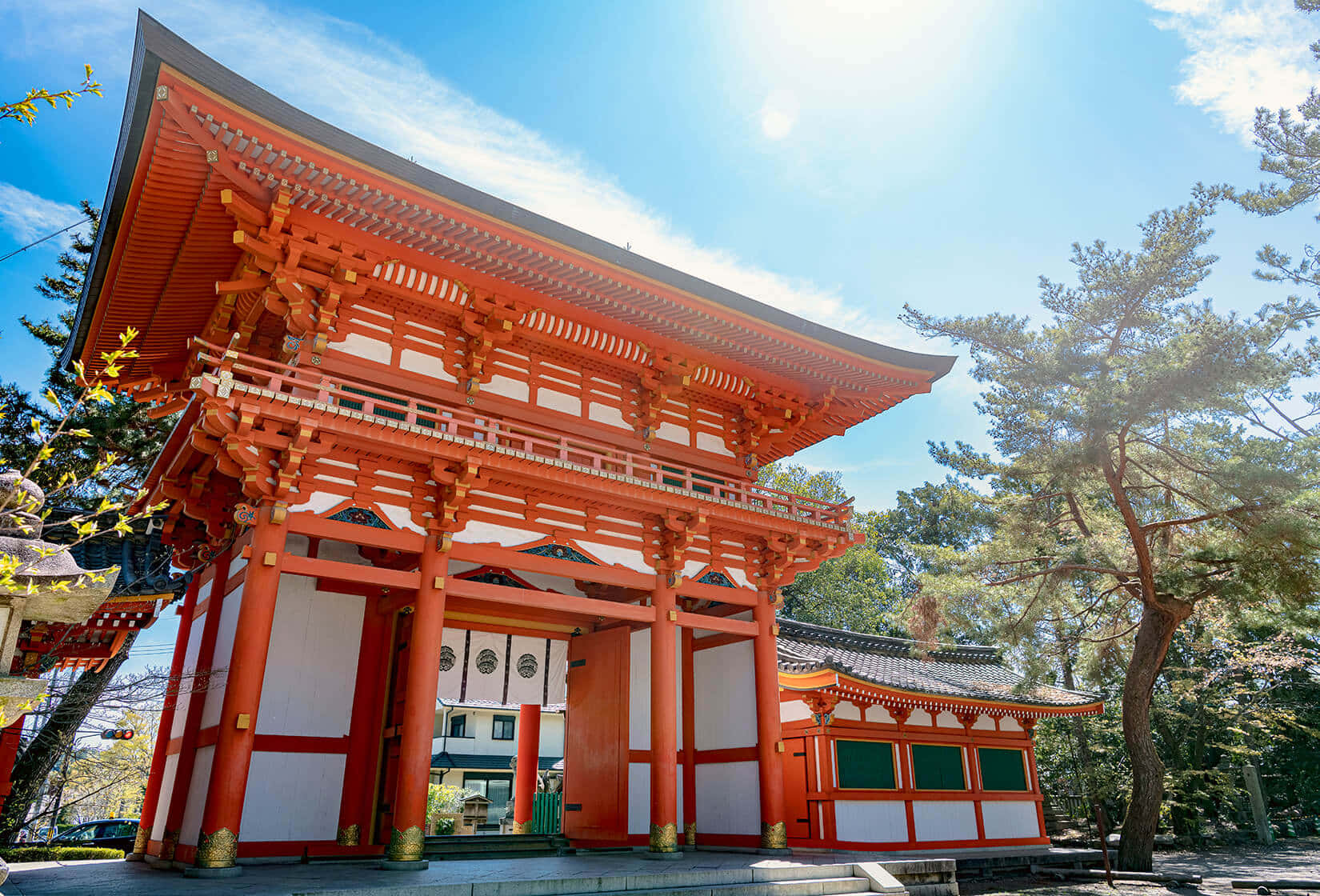 日式 桌布 壁紙 — 一座鮮紅色的傳統日本寺廟大門在明亮的藍天下高聳，陽光穿過周圍的松樹灑落。