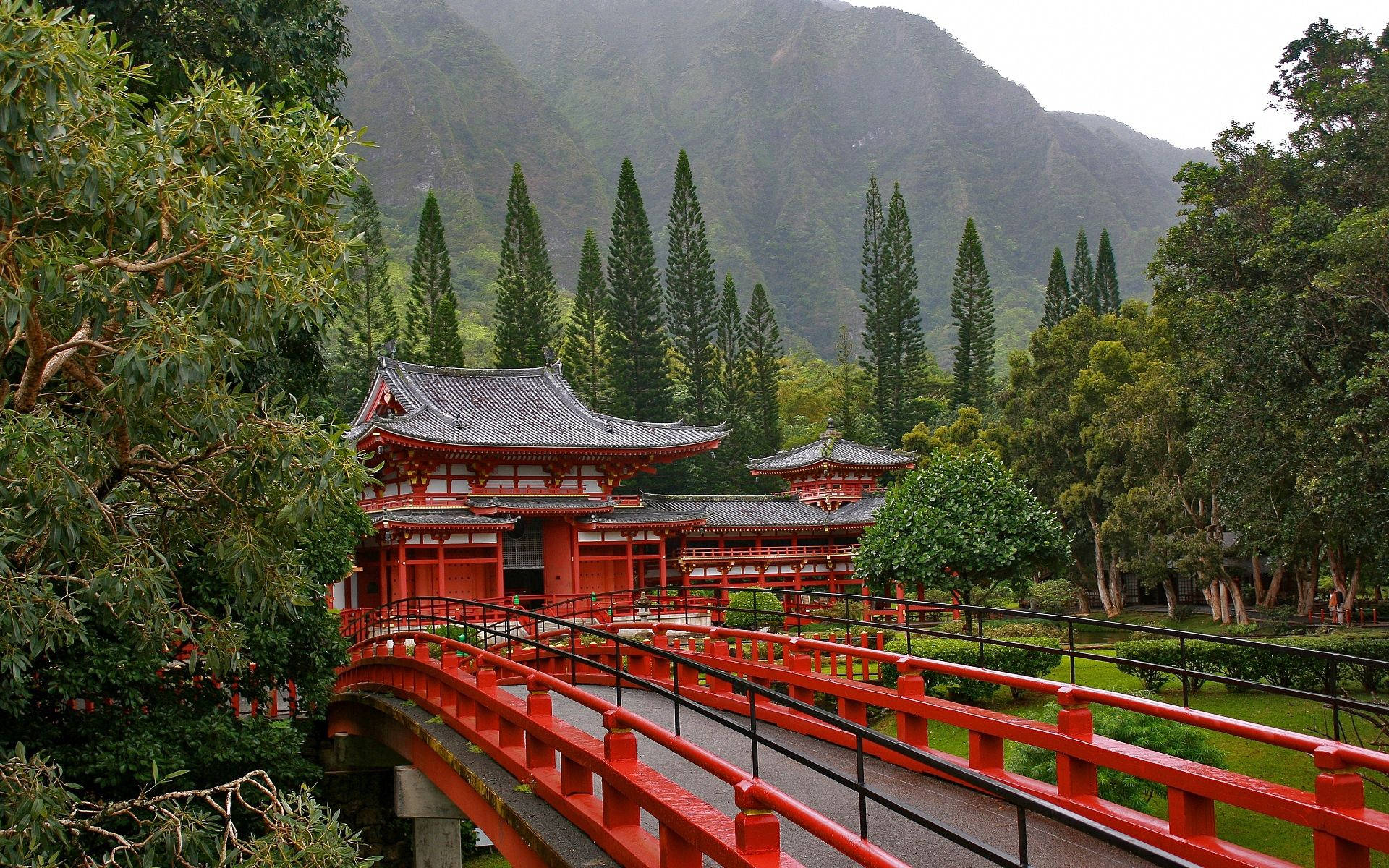 日本 桌布 壁紙 — 一座鮮紅色的日本寺廟橋樑拱跨在溪流之上，周圍環繞著茂密的綠樹和霧氣繚繞的山巒。