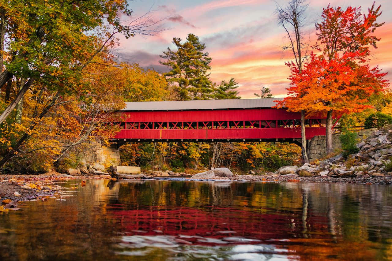 Fall Bridge 桌布 壁紙 — 一座鮮紅色的有蓋橋樑橫跨在日落時分的平靜河流上，周圍的秋葉倒映在水中。
