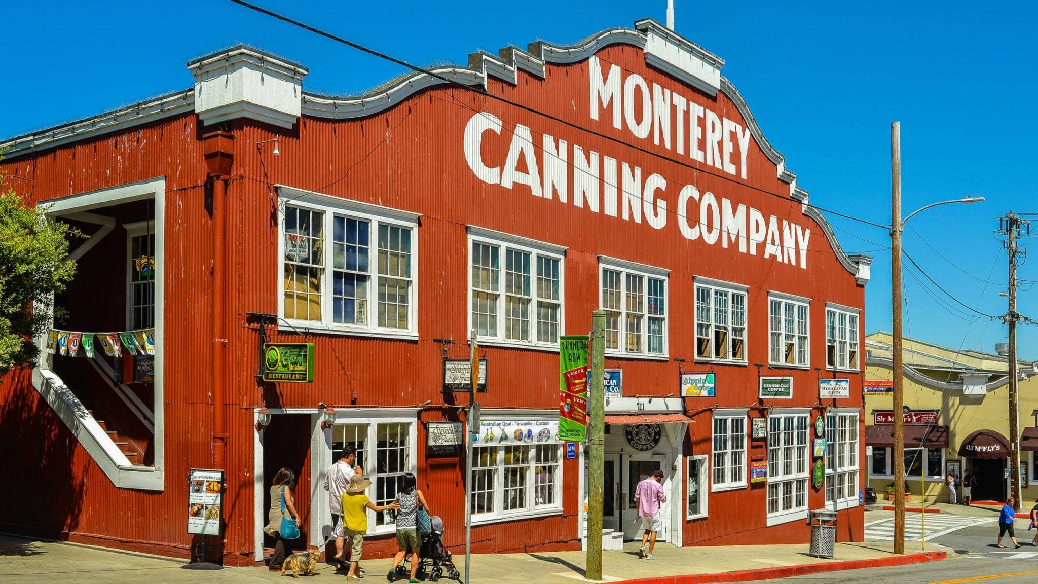Building 桌布 壁紙 — 一座鮮紅色的歷史建築 Monterey Canning Company，帶有白色飾邊，人們在晴朗的藍天下走在人行道上。